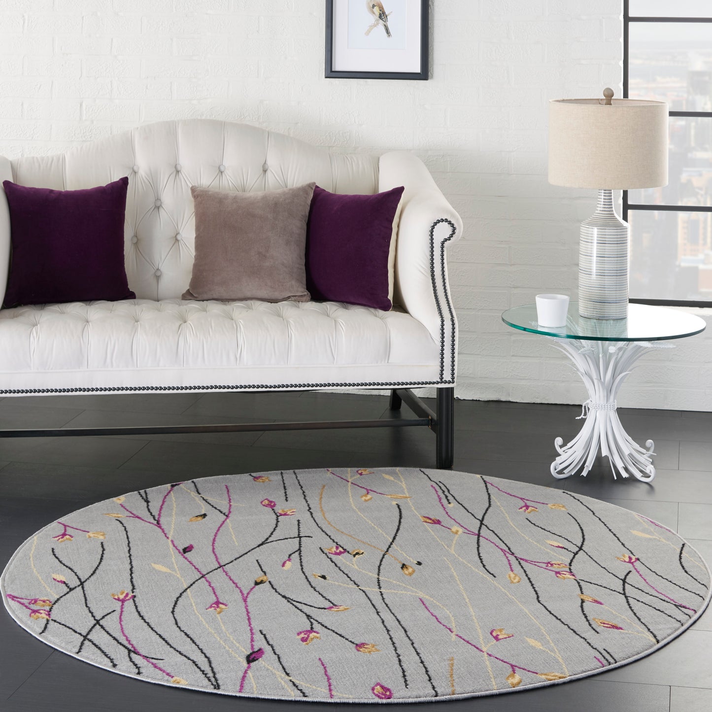 White tufted sofa with purple and taupe cushions, gray abstract rug with colorful branches, glass side table, beige lamp.