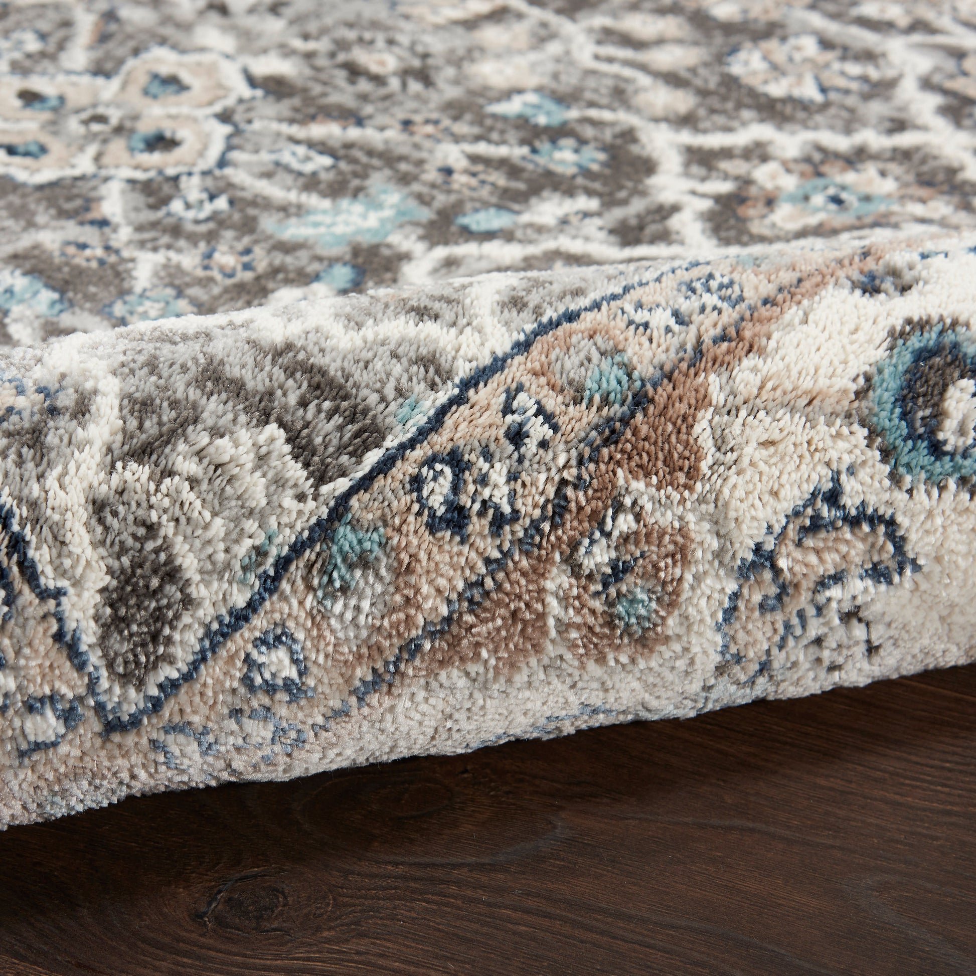 Close-up of a traditional beige-toned rug with blue and teal accents and an ornate border, soft pile on dark wood floor.