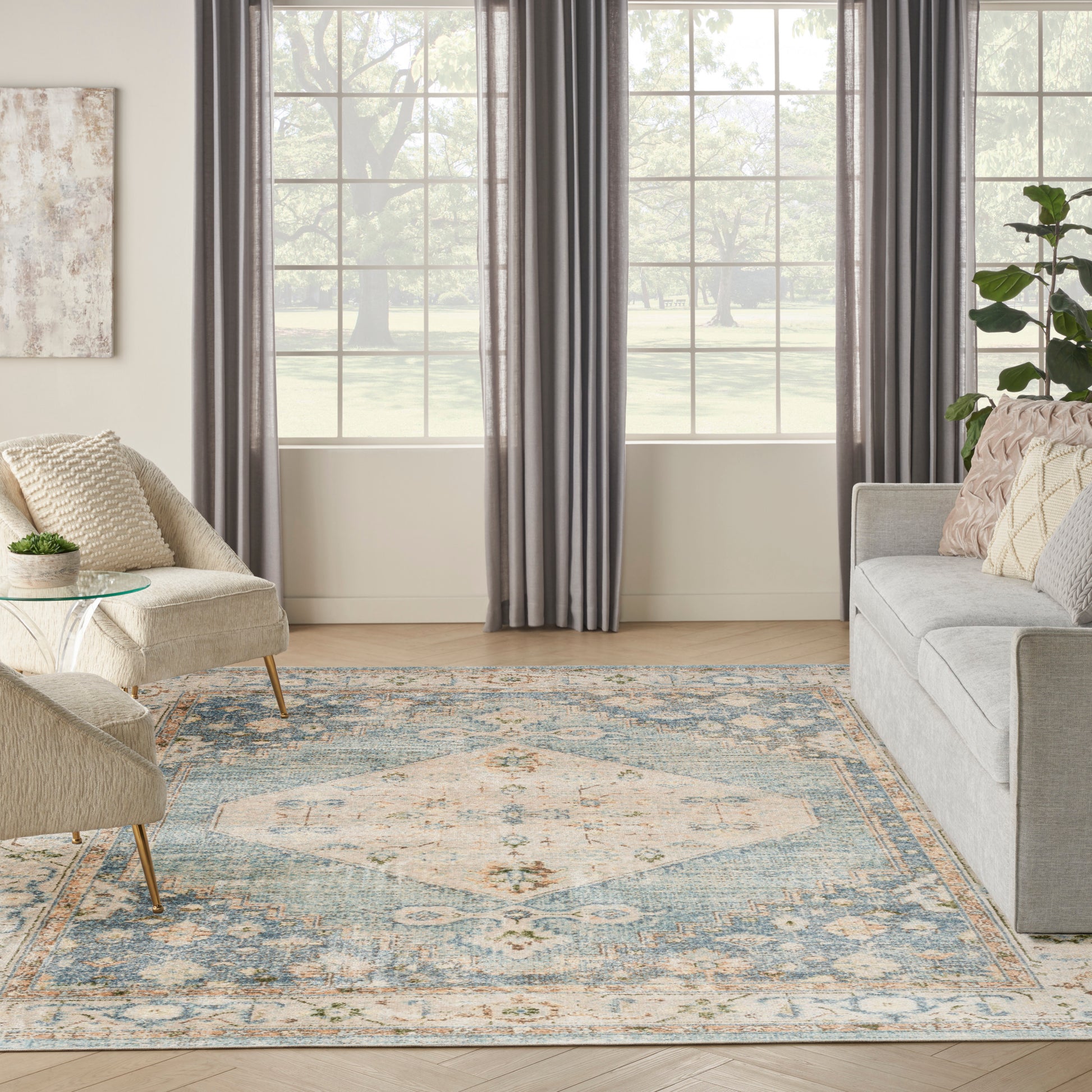 Living room with blue-and-beige ornate area rug, gray sofa, cream accent chair, glass side table, charcoal curtains and a plant.