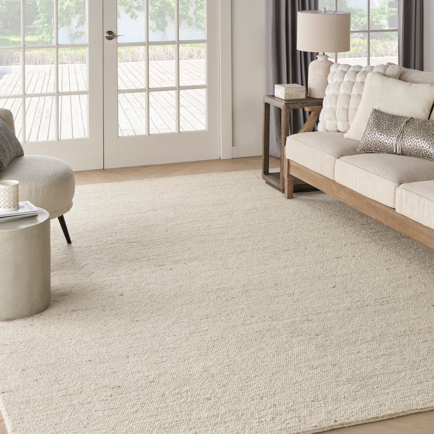 Neutral living room with large beige woven rug, tufted beige sofa, round ottoman, wood side table, lamp, and French doors.