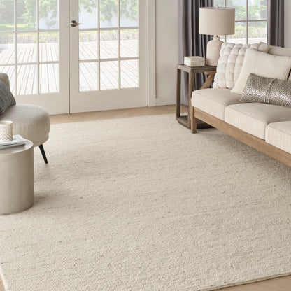 Neutral living room with large beige woven rug, tufted beige sofa, round ottoman, wood side table, lamp, and French doors.