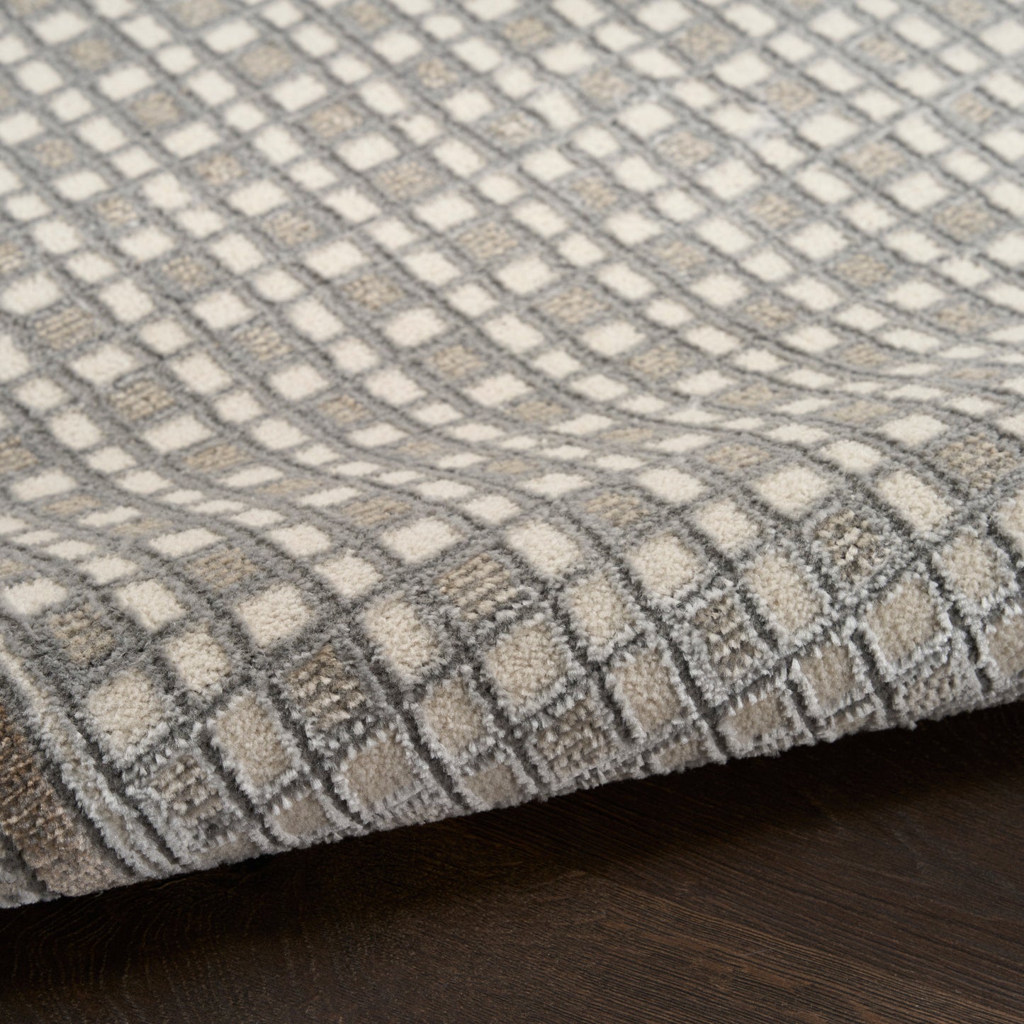 Soft area rug with gray & ivory checker grid pattern, plush textured pile, folded edge on dark wood floor.