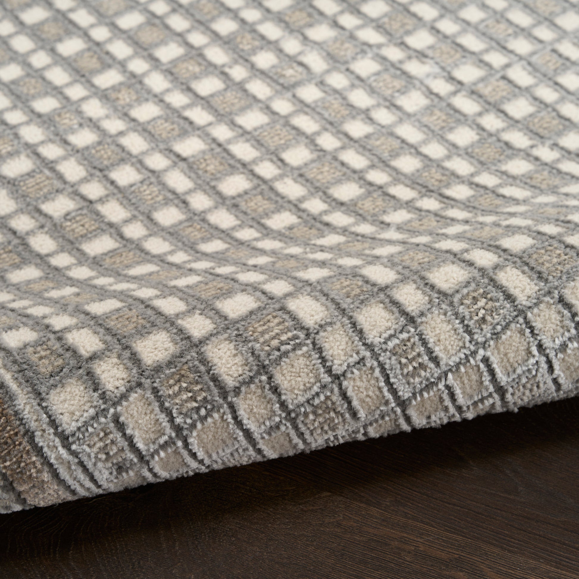 Soft area rug with gray & ivory checker grid pattern, plush textured pile, folded edge on dark wood floor.