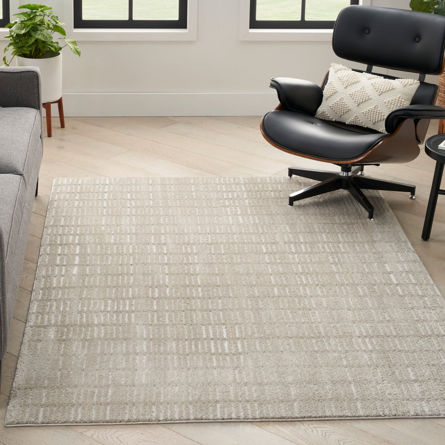 Beige textured rug with subtle grid on light wood floor beside black leather lounge chair and white pillow.