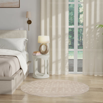 Neutral bedroom with cream bedding, round white side table, glass lamp, stack of books, beige circular rug, sheer curtains.