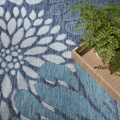 Blue woven area rug with white and navy leaf motifs; natural fiber rectangular tray with a potted green fern.