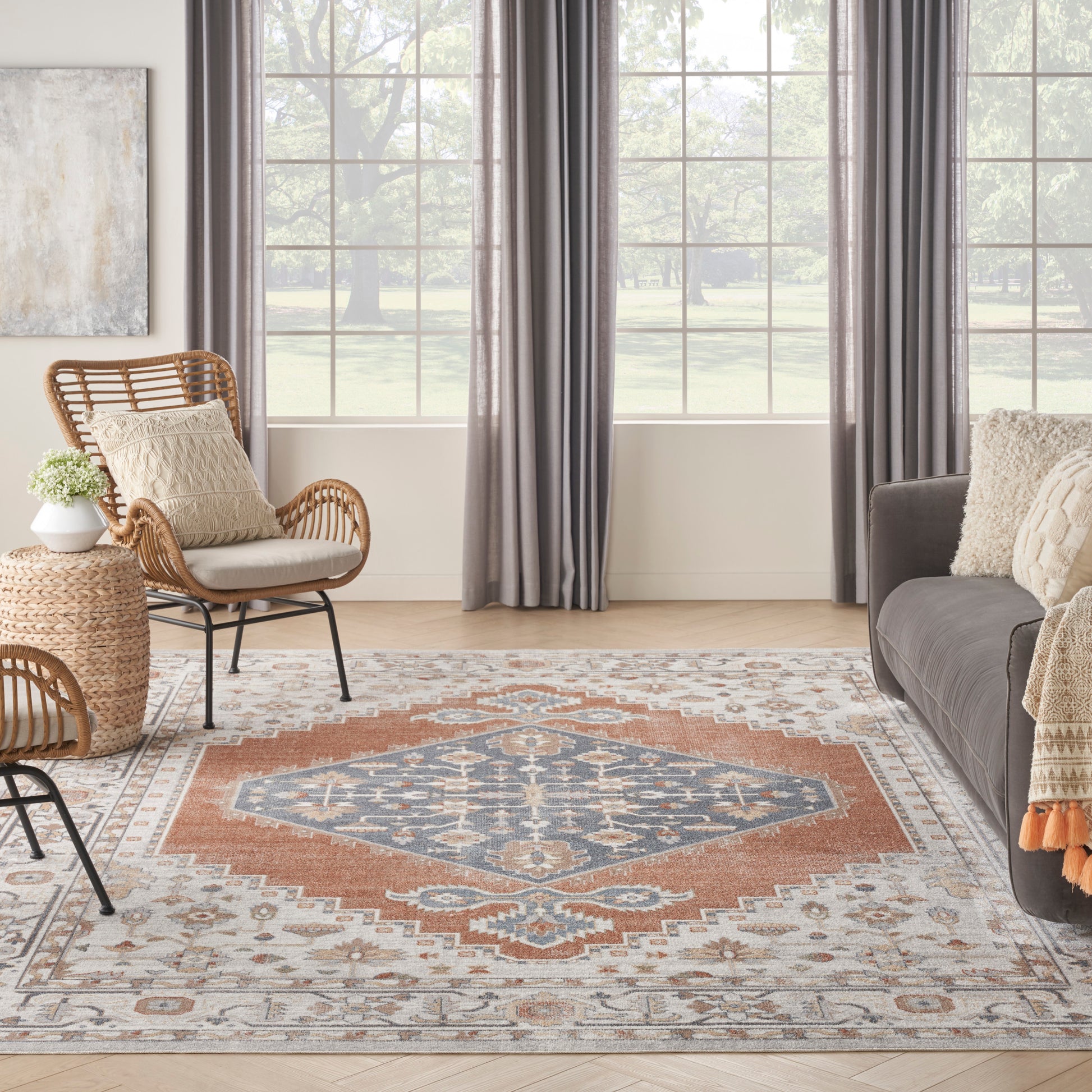 Living room with blue and orange patterned area rug, wicker chair and round side table, gray sofa, and large windows with gray drapes.