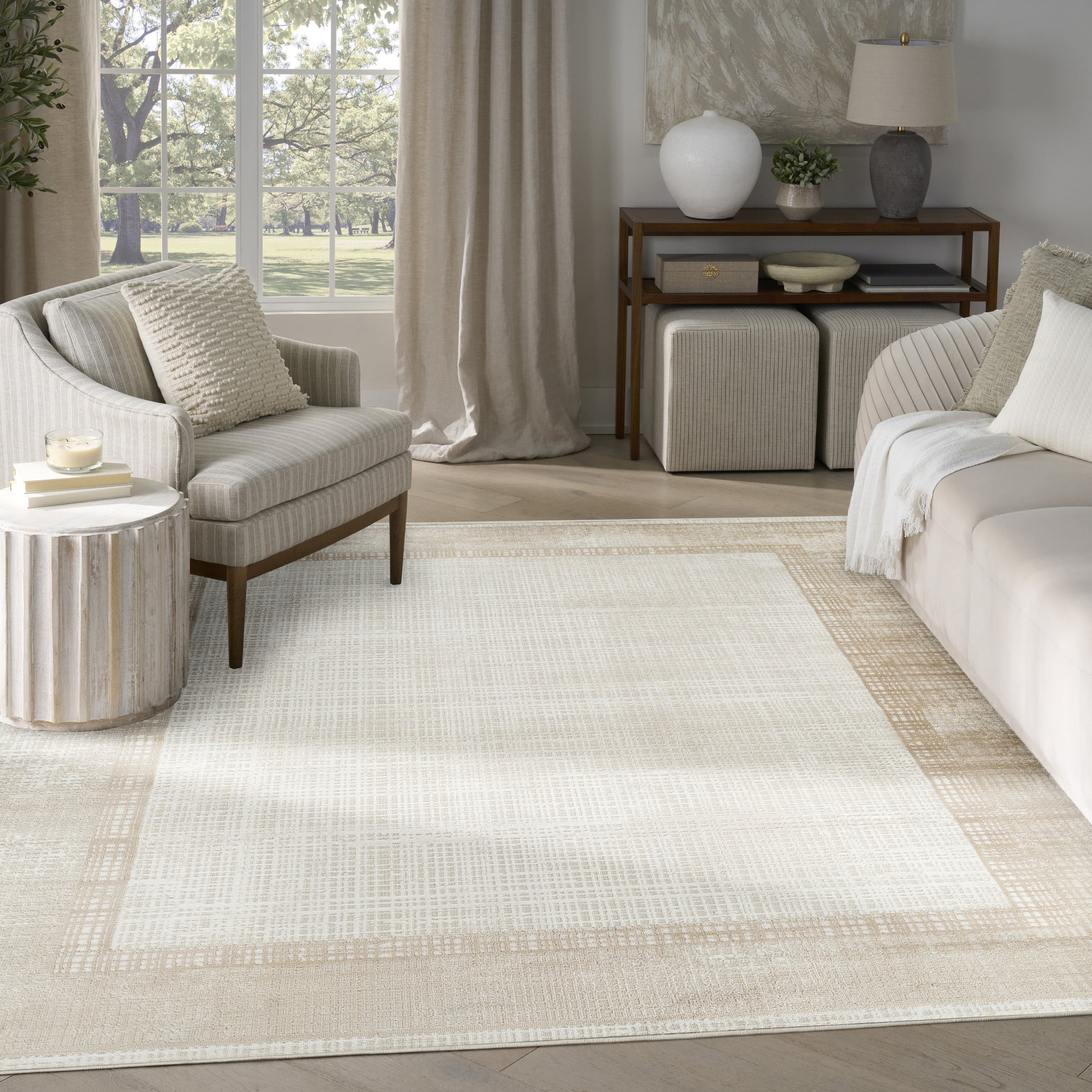 Neutral living room with cream striped armchair, beige sofa, woven bordered rug, wood console and ottomans near a window.