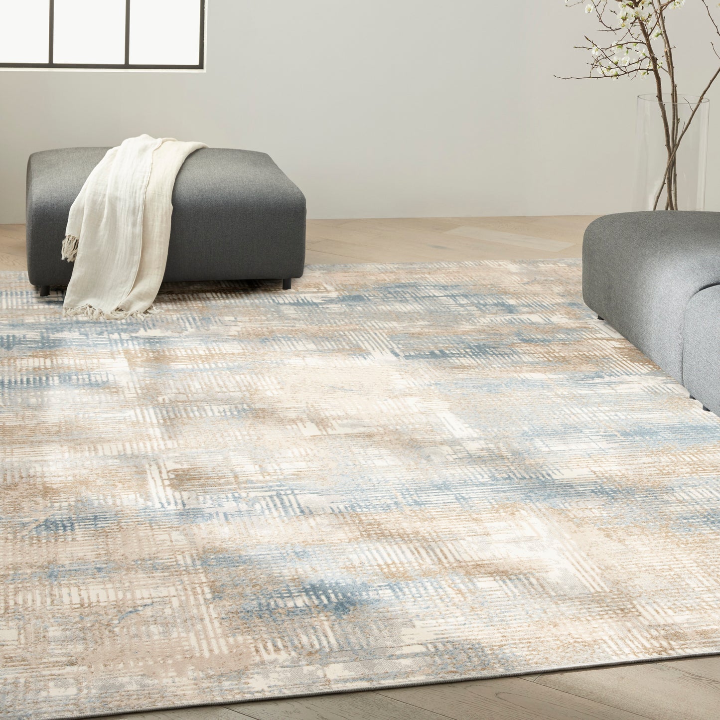 Modern abstract beige, blue and cream area rug with gray ottomans and a white throw blanket.