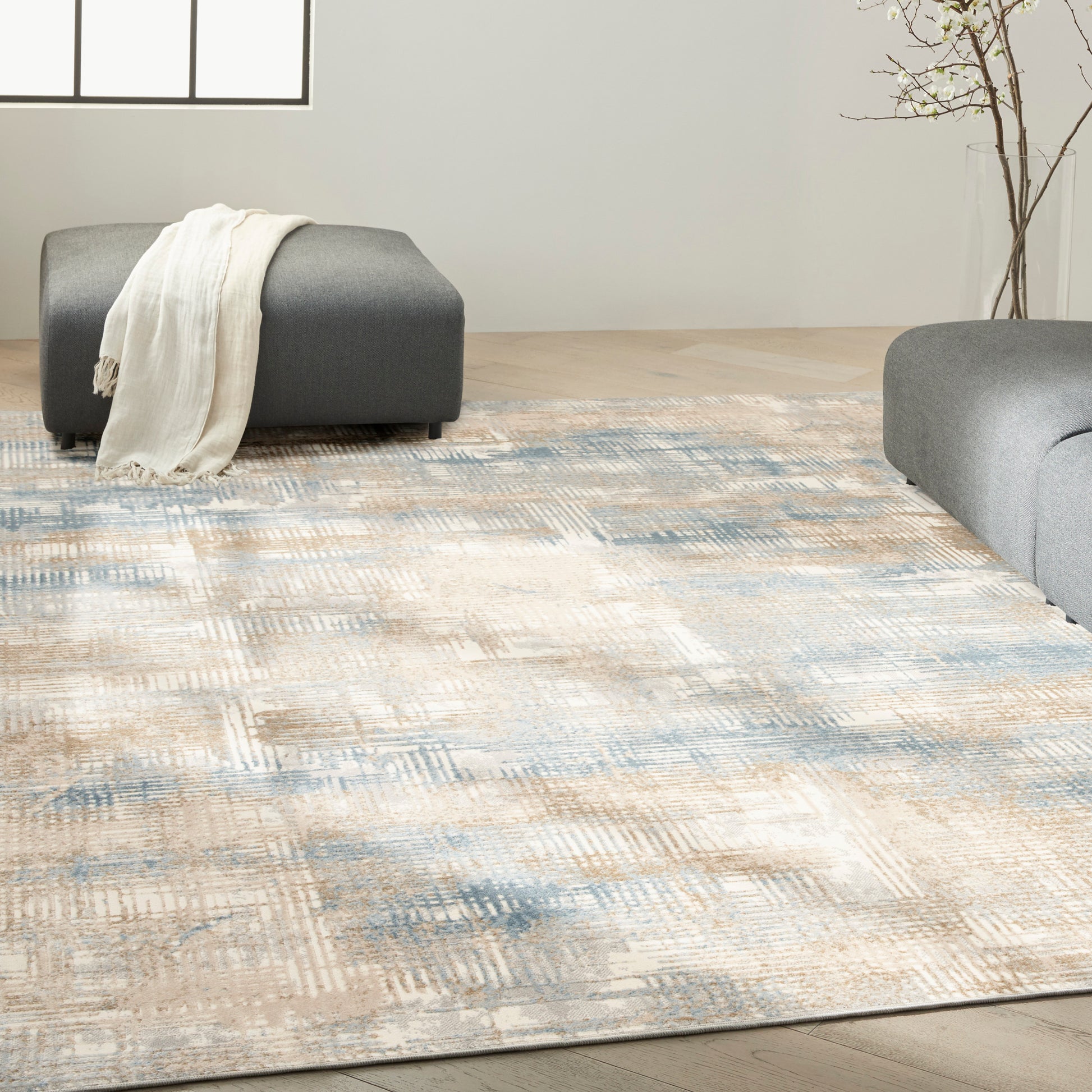 Modern abstract beige, blue and cream area rug with gray ottomans and a white throw blanket.
