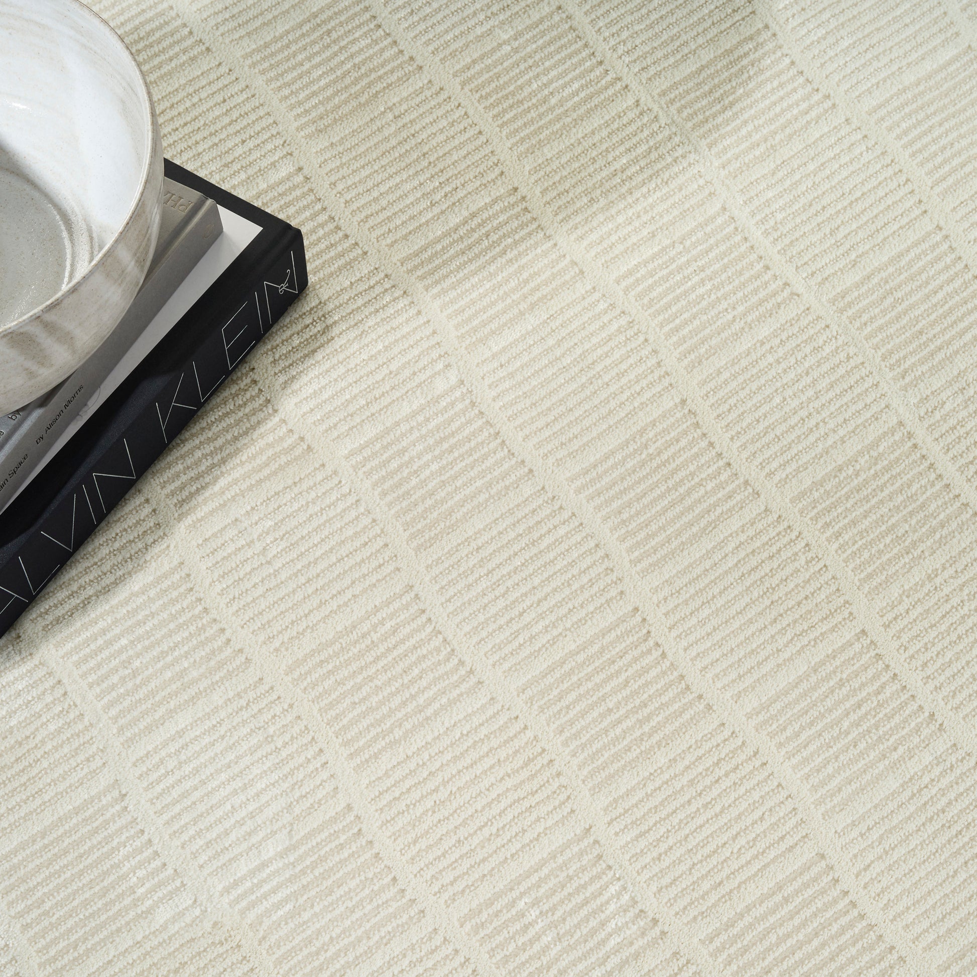 Cream textured rug with square grid pattern; white ceramic cup on a dark rectangular book.