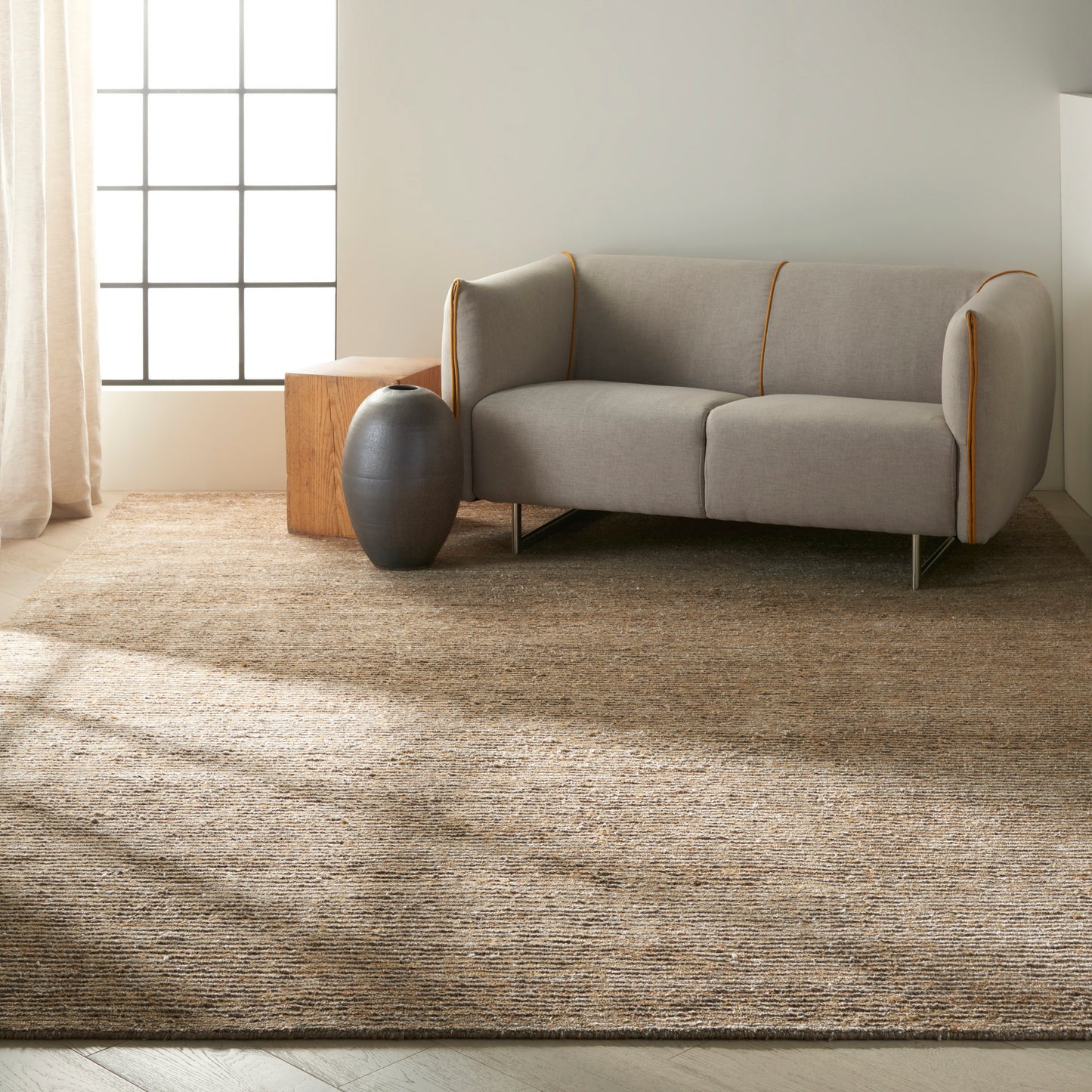 Light gray sofa with orange piping on a textured tan rug, beside a dark ceramic vase and a wooden cube side table.