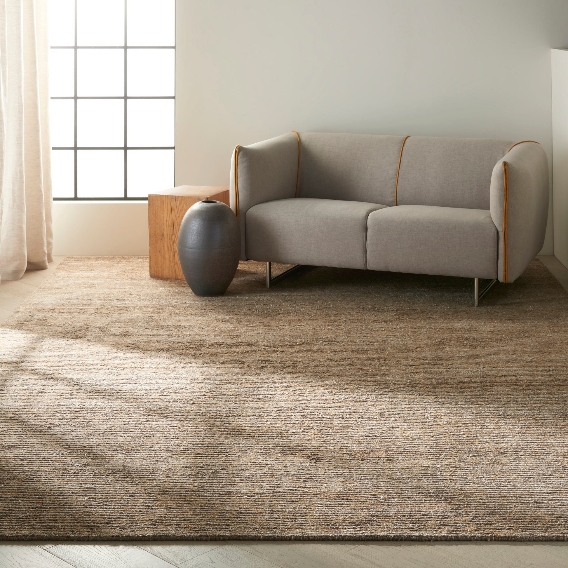 Light gray sofa with orange piping on a textured tan rug, beside a dark ceramic vase and a wooden cube side table.