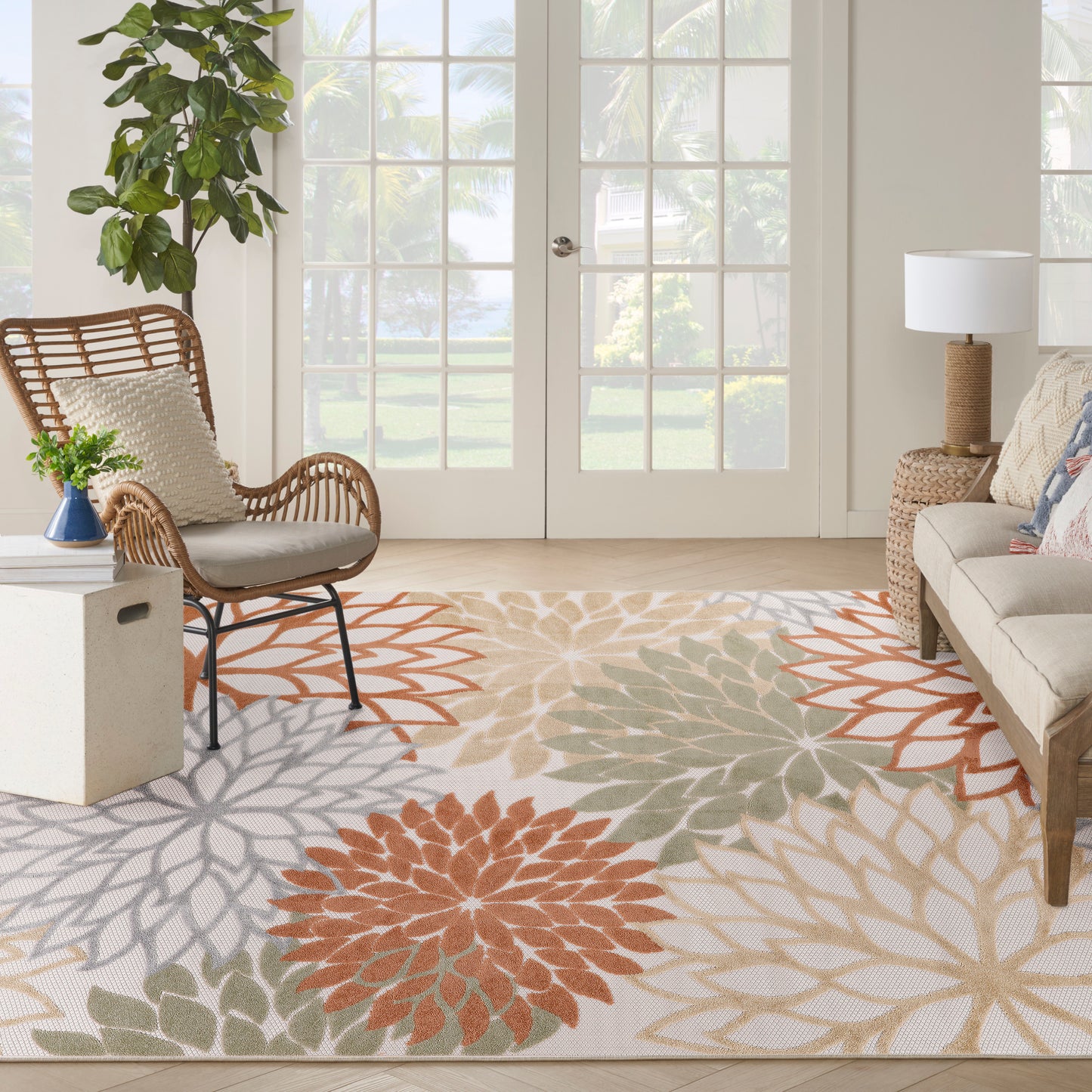 Sunlit living room with an outdoor floral-patterned rug in orange, gray, and sage; wicker chair, beige sofa, potted plant.