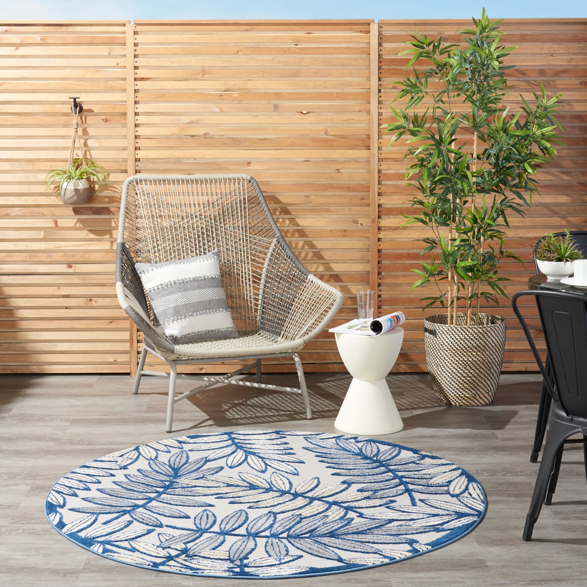 Round blue leaf-pattern outdoor rug beside a woven lounge chair, white side table, and potted plant by a wooden slat fence.