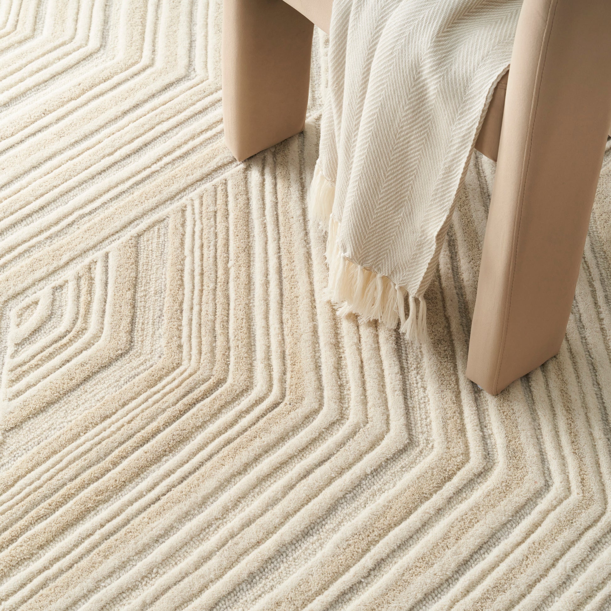 Cream textured rug with raised chevron pattern under a beige chair and fringed cream throw.