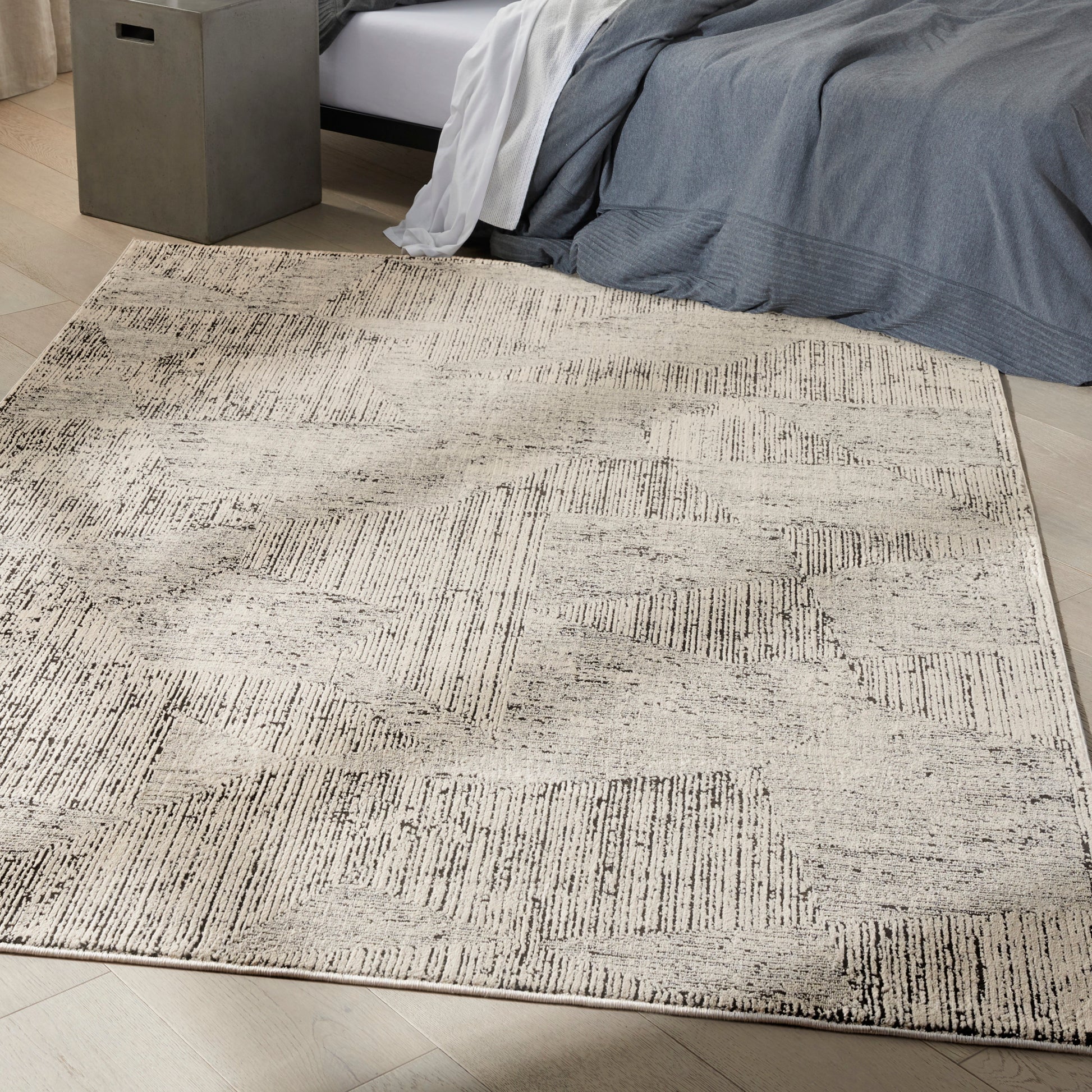 Cream-gray abstract-pattern area rug on light tile floor beside a bed with blue-gray bedding and a gray storage ottoman.