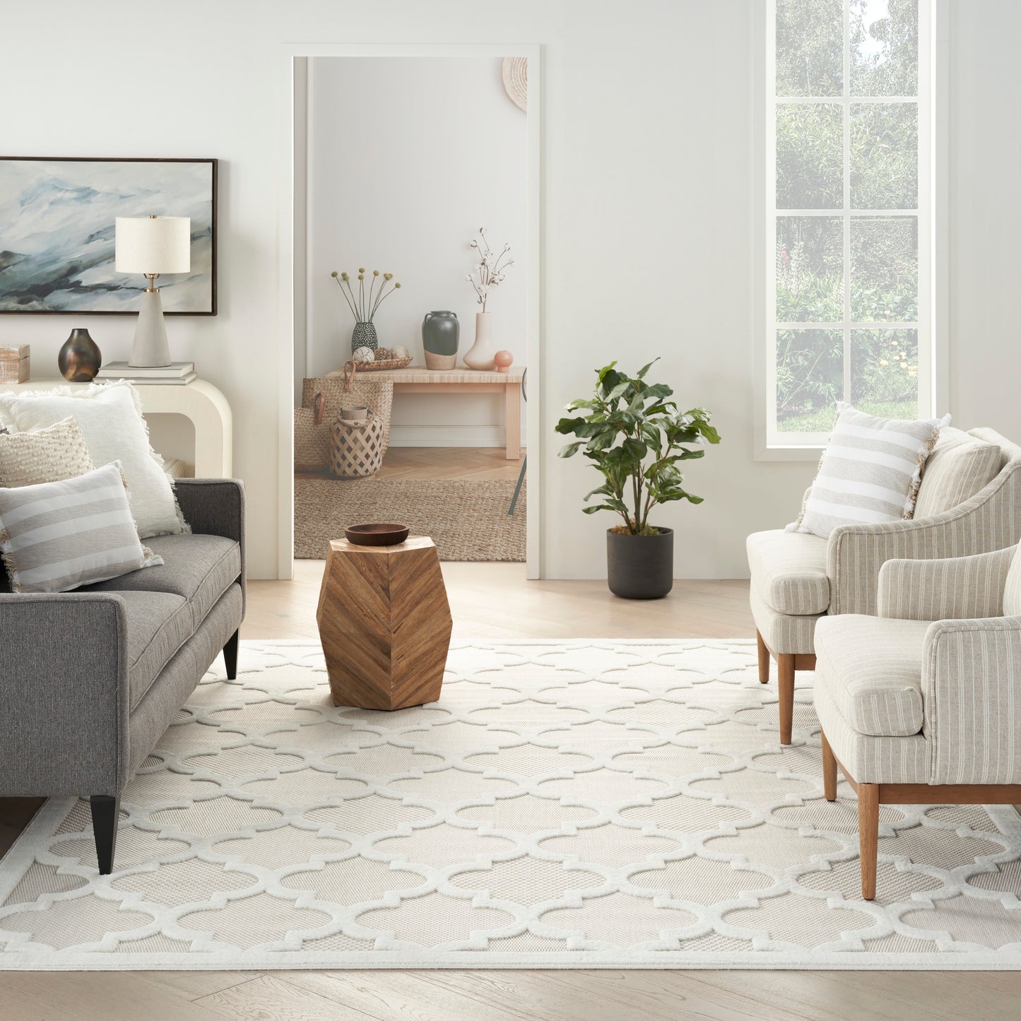 Bright modern living room with a beige geometric rug, gray sofa, cream striped armchairs, faceted wooden stool, potted plant, and a large window.