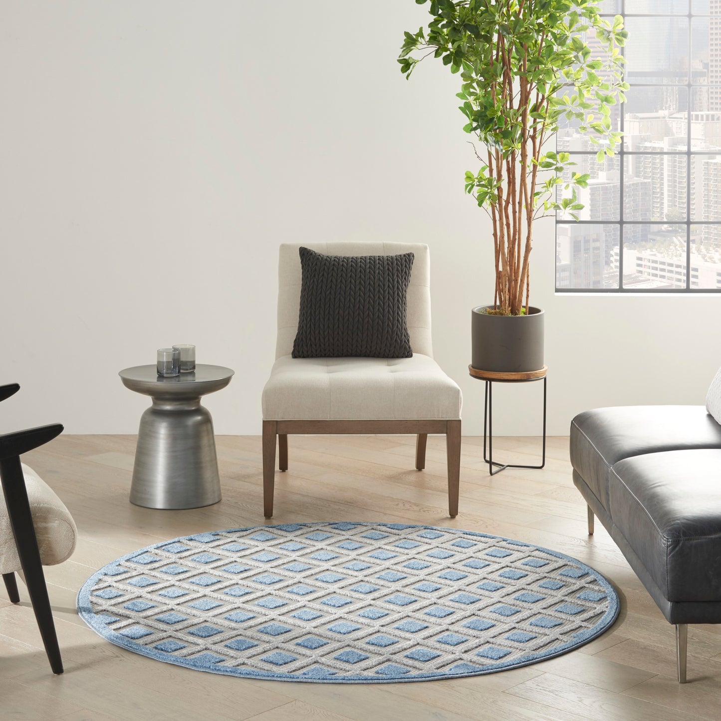 Round blue geometric rug in a living room with cream chair, black pillow, gray side table, leather couch, and tall plant.