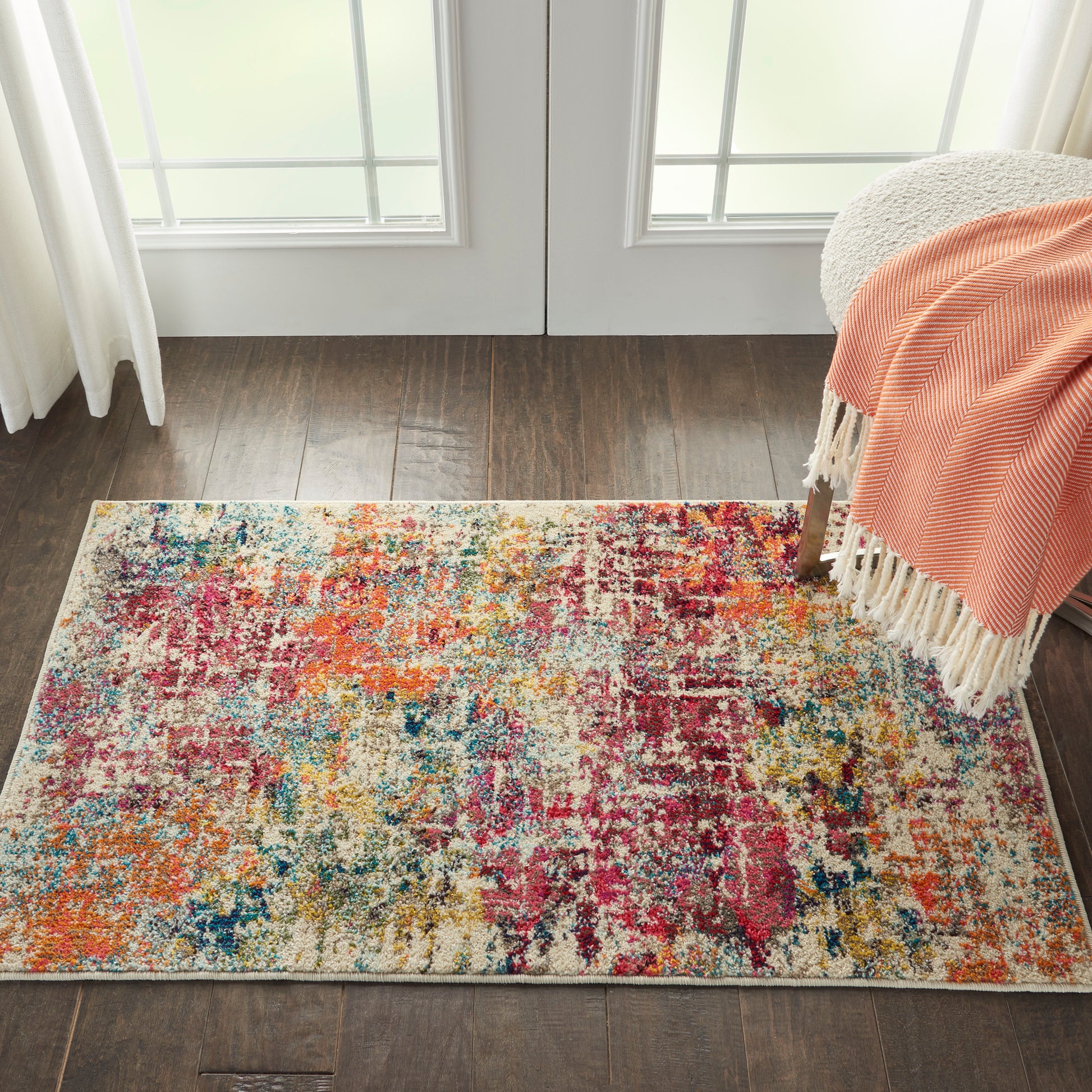 Rectangular abstract multicolor area rug on dark hardwood floor, beside a chair with an orange fringed throw.