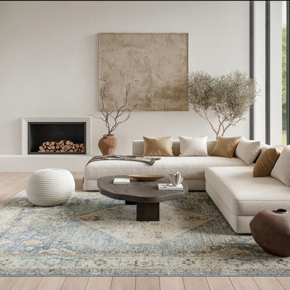 Neutral living room with beige sectional, dark round wood coffee table, white knit pouf, blue patterned rug, and fireplace.