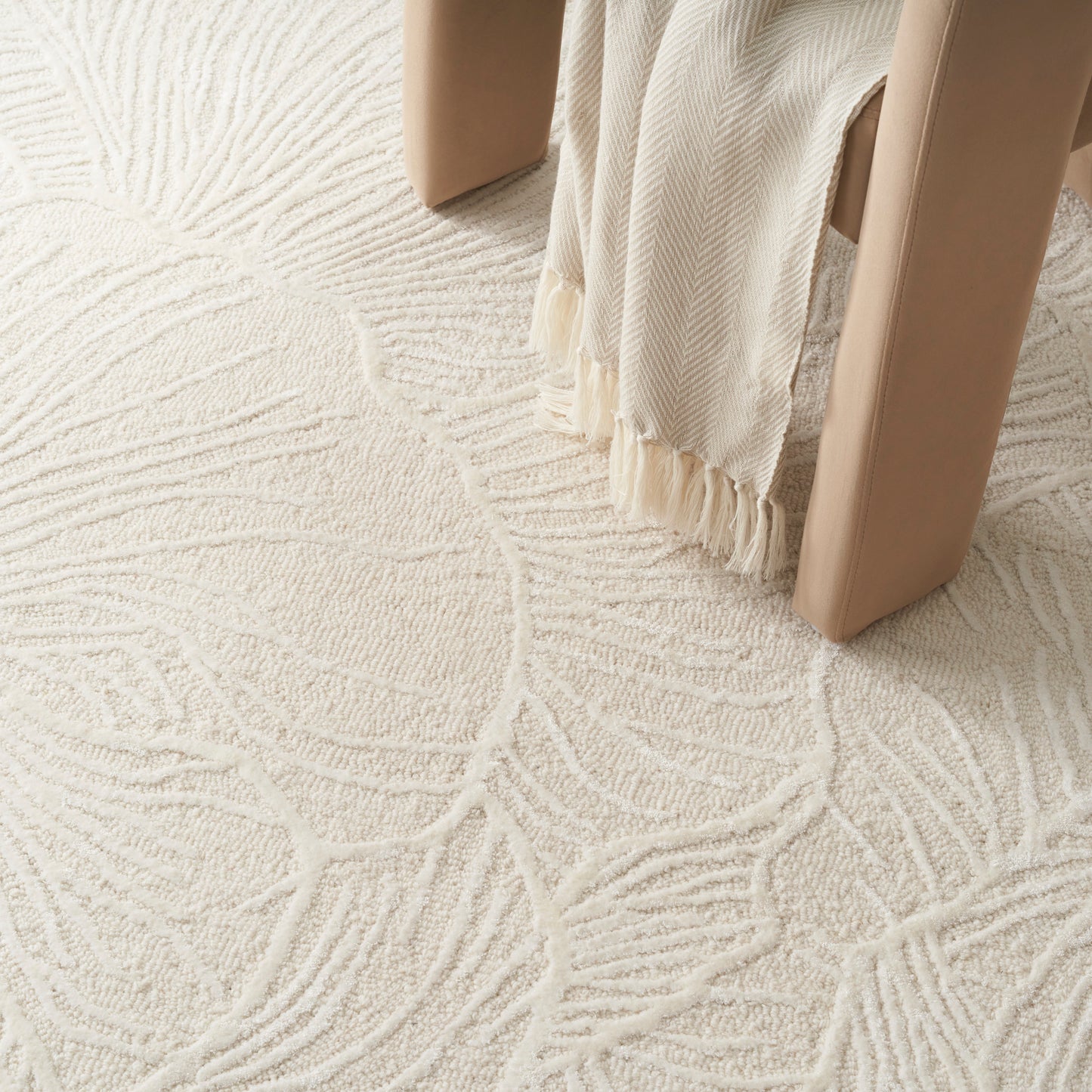 Cream textured rug with an abstract leaf pattern, beige furniture legs, and fringe-draped fabric.