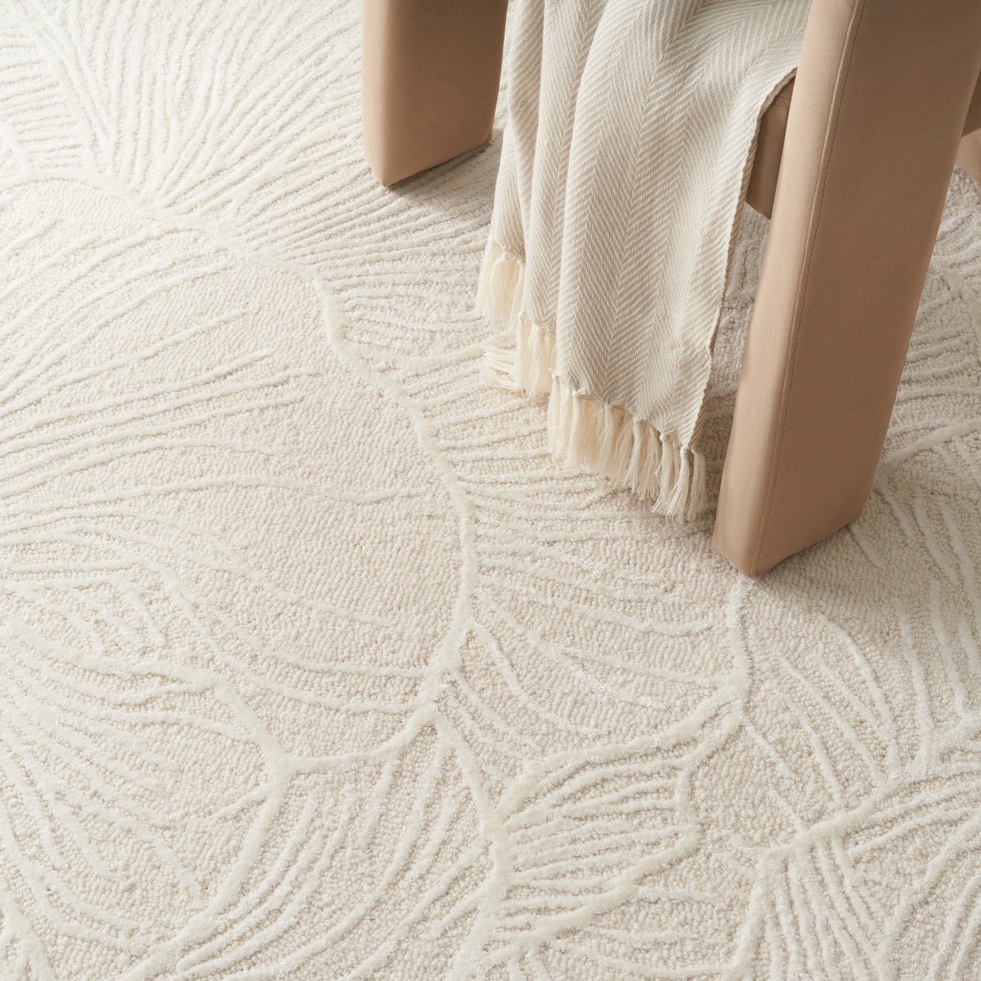 Cream textured rug with an abstract leaf pattern, beige furniture legs, and fringe-draped fabric.