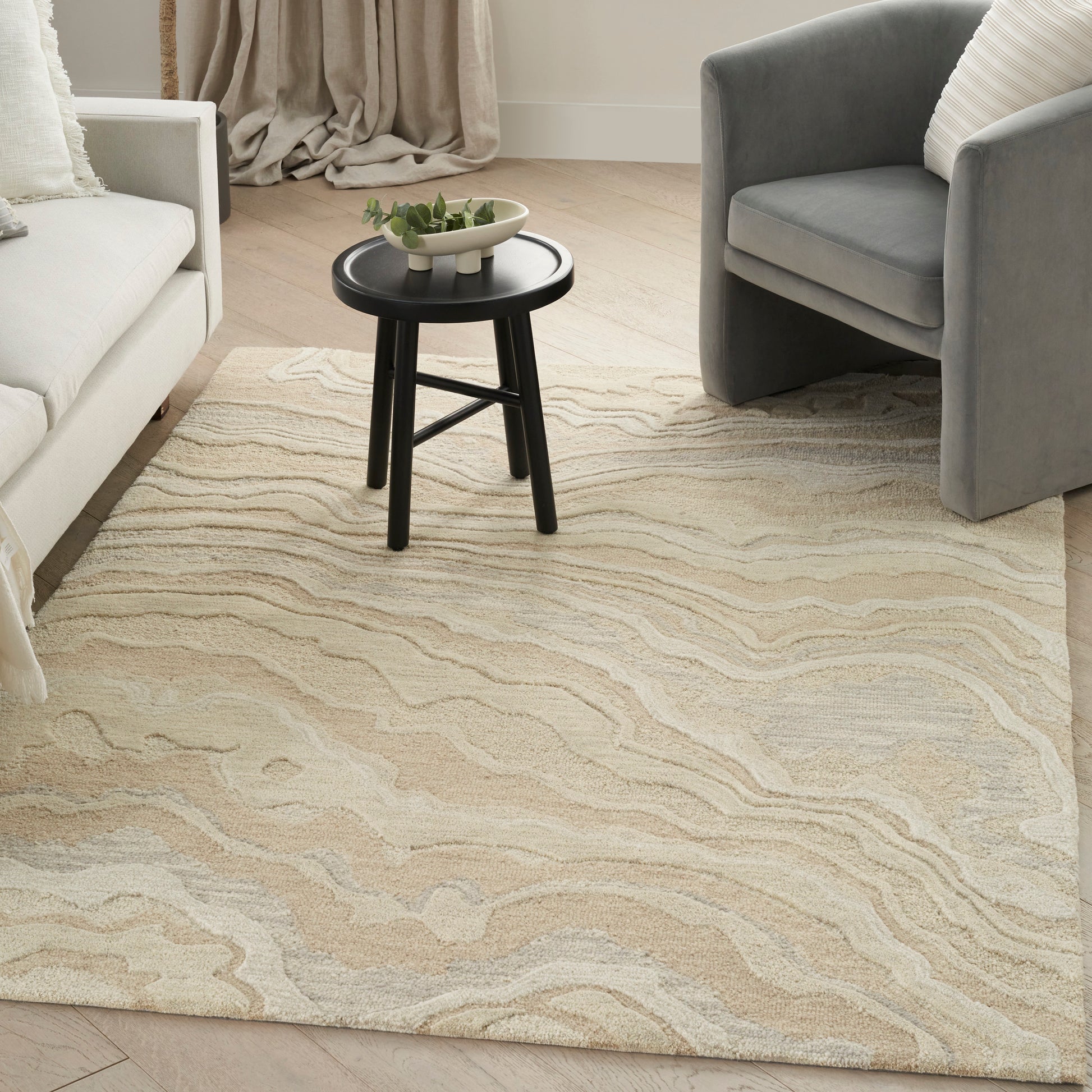 Beige wavy-pattern area rug between a white sofa and gray armchair, small round black side table with a plant.