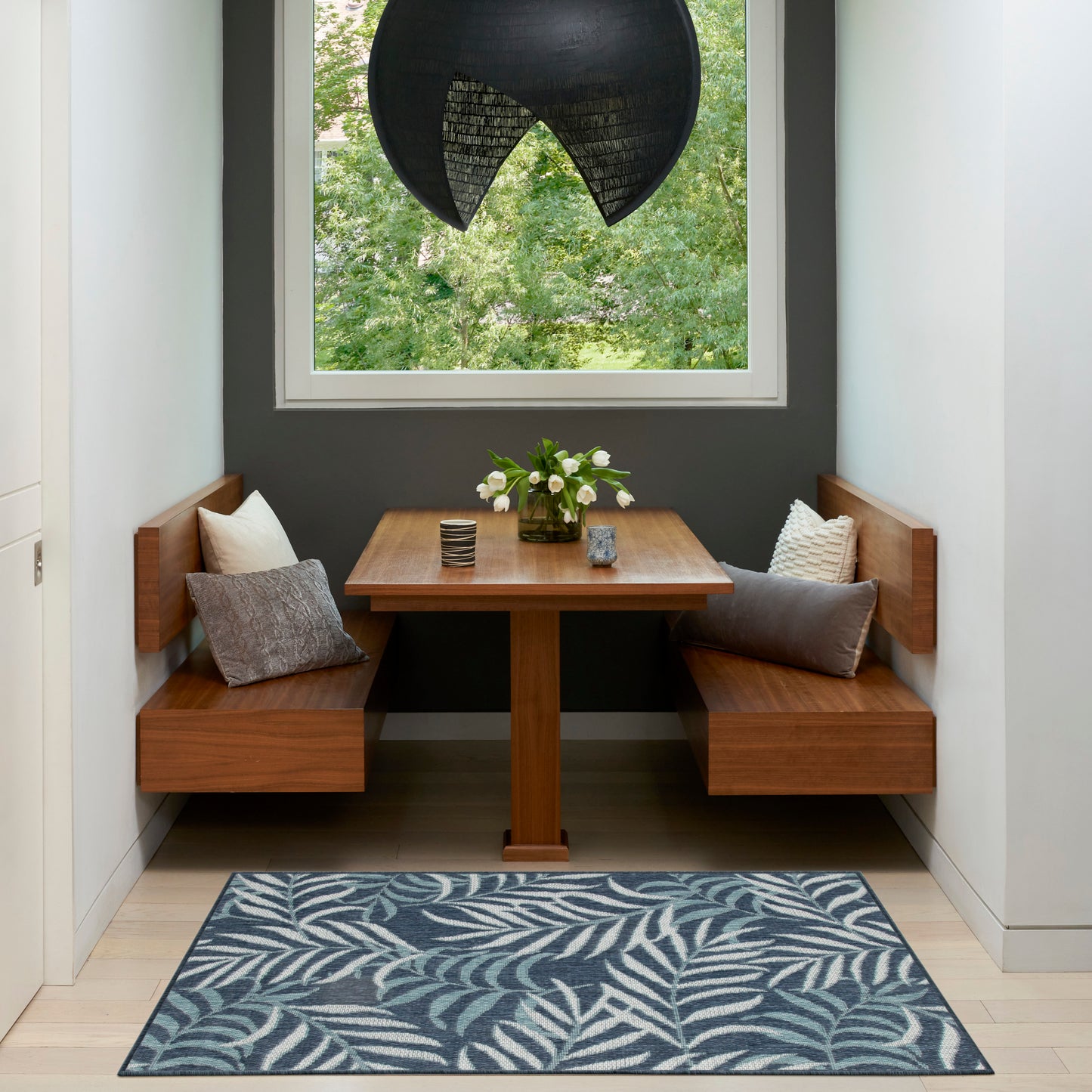 Walnut built-in banquette with cushions around a rectangular table, dark wall, large window, black pendant light, & blue leaf rug.