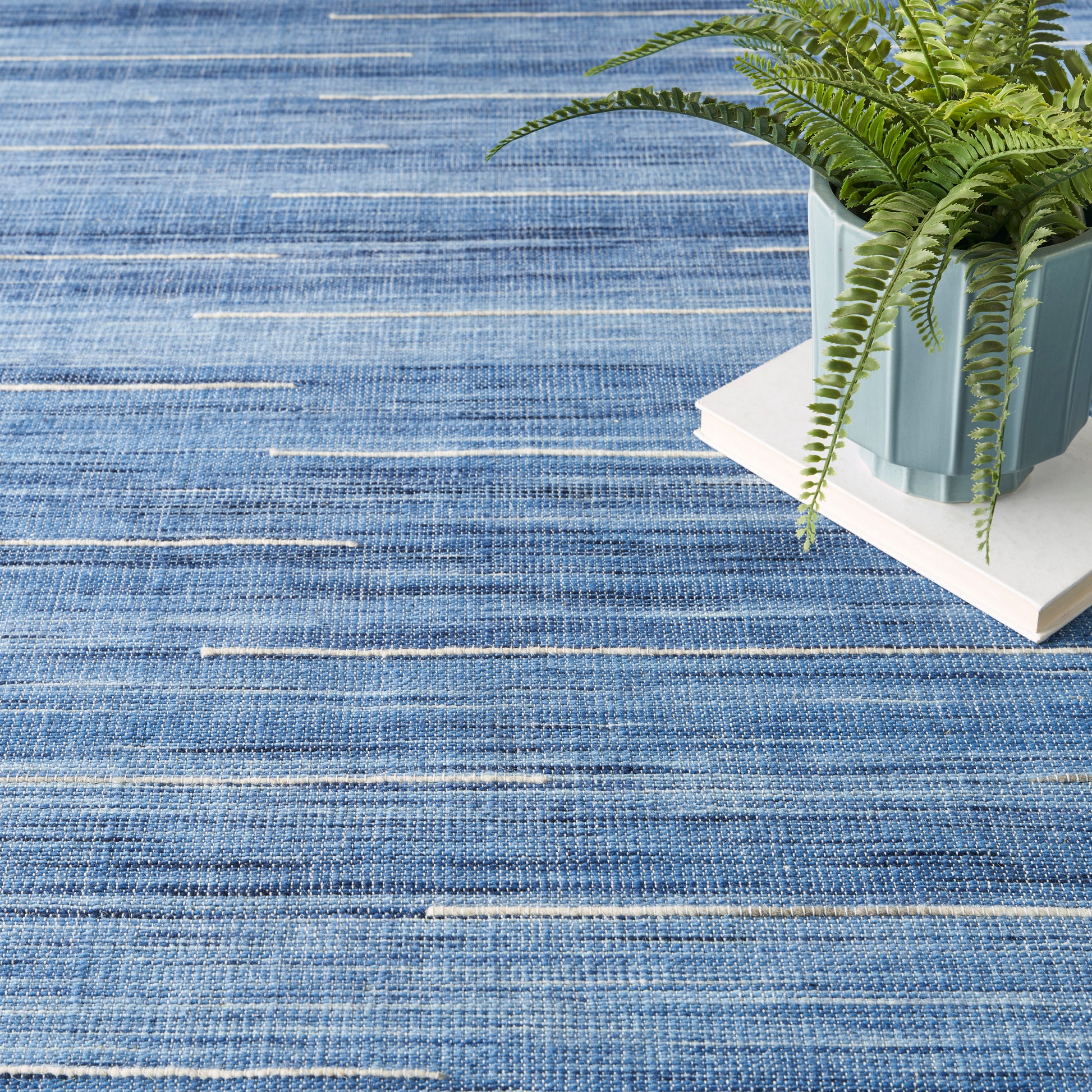 Blue striped woven rug with white accents, featuring a potted fern in a blue planter resting on white books.