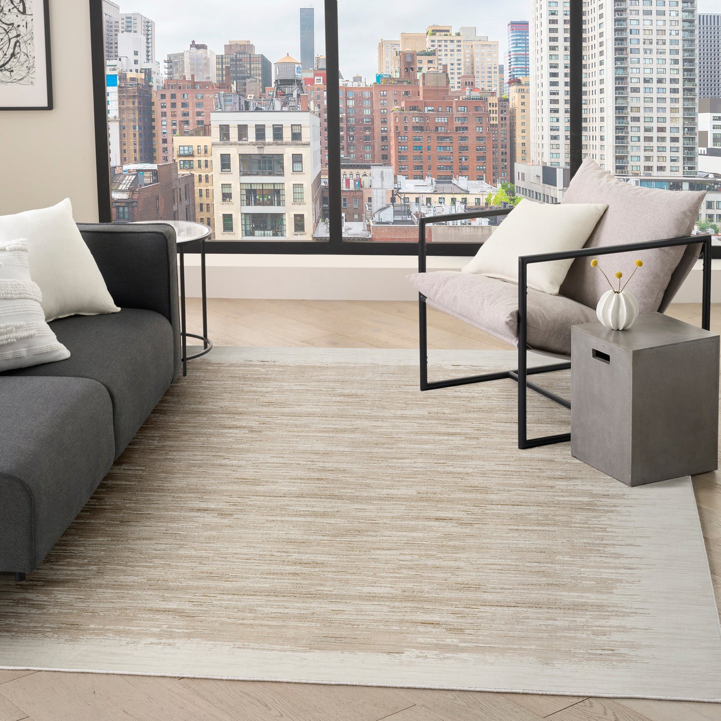Neutral living room with city view, gray sofa, beige cushions, black metal chair, concrete side table, white vase & cream rug