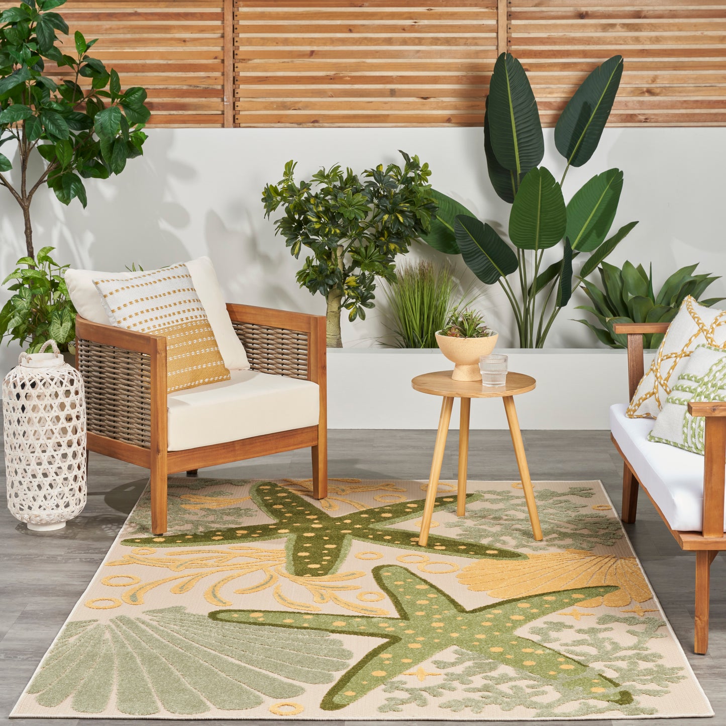 Outdoor patio with wooden-framed chairs, white cushions, a green tropical rug, round wooden table, and lush potted plants.