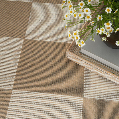 Woven checkerboard floor mat in brown and beige with a wicker tray and a potted daisy arrangement atop a stack of books.