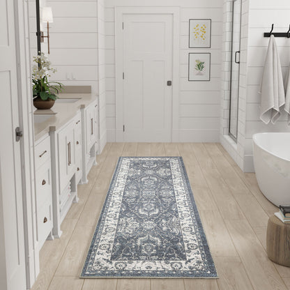 Blue-gray ornate bathroom runner rug on light wood floor beside white vanity, plant, towels, and tub.