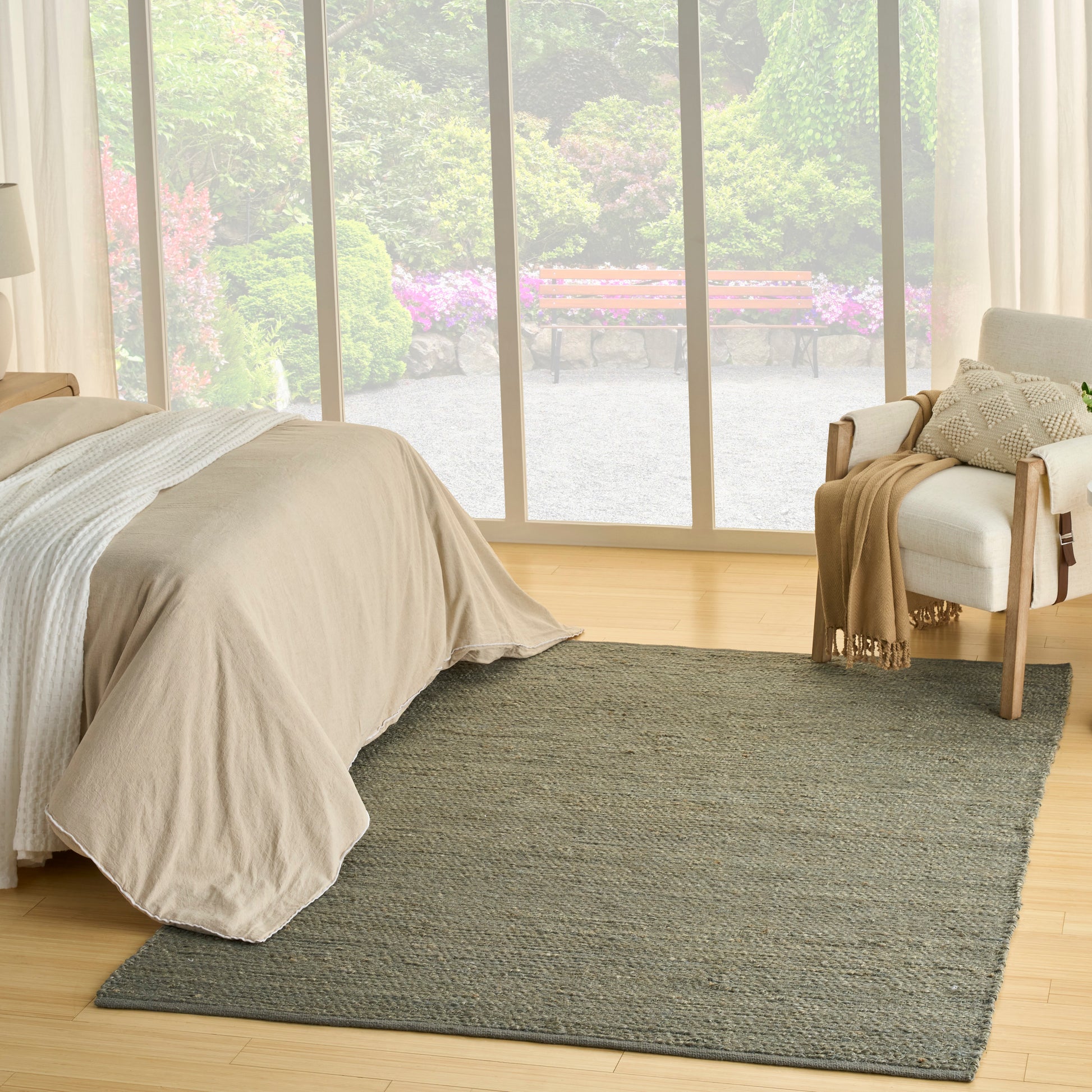 Neutral-toned bedroom with beige duvet, light wood floor, large grey rug, floor-to-ceiling windows; cream chair with throw.