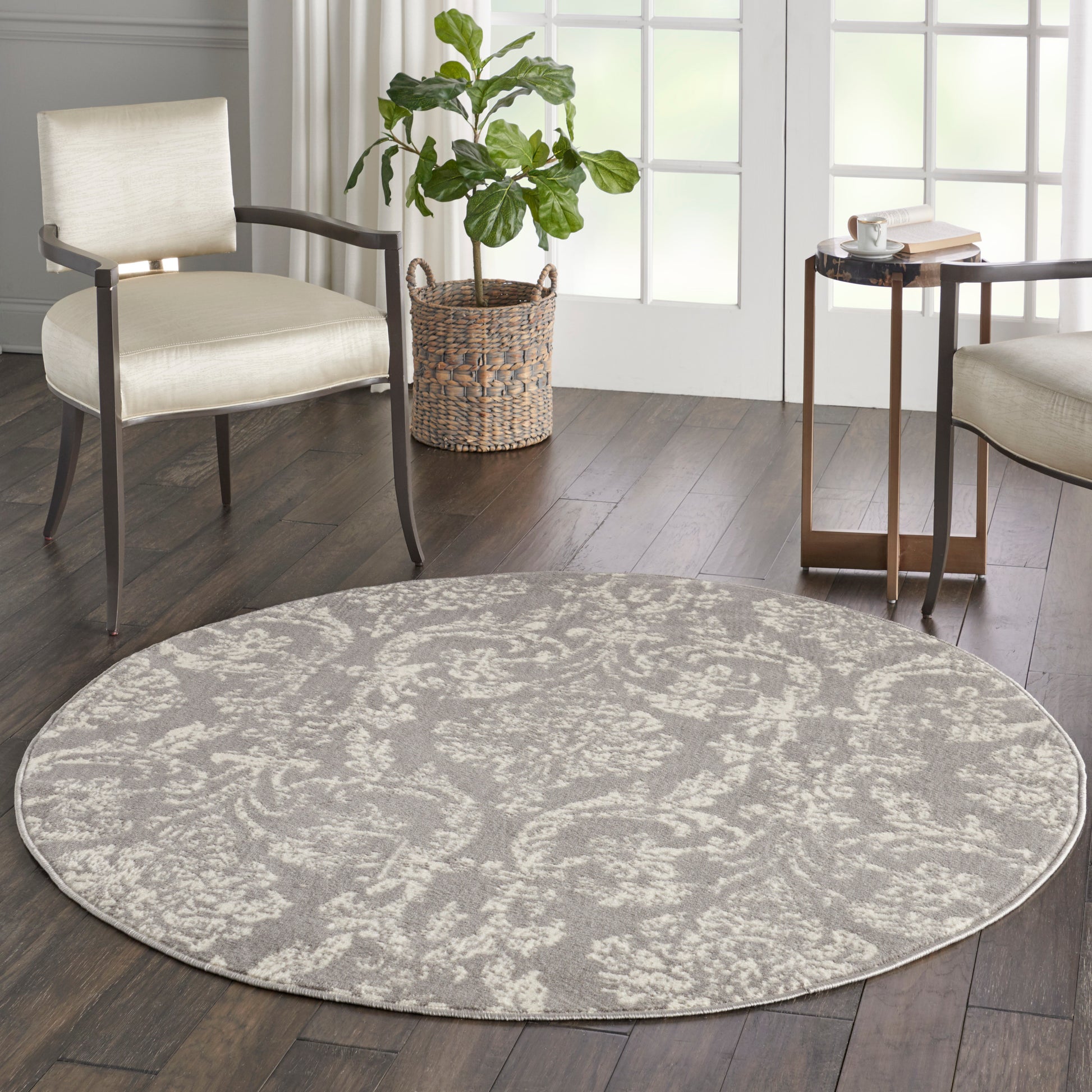 Round gray rug with ivory floral pattern on dark hardwood flooring; beige upholstered chairs and a wicker plant basket.