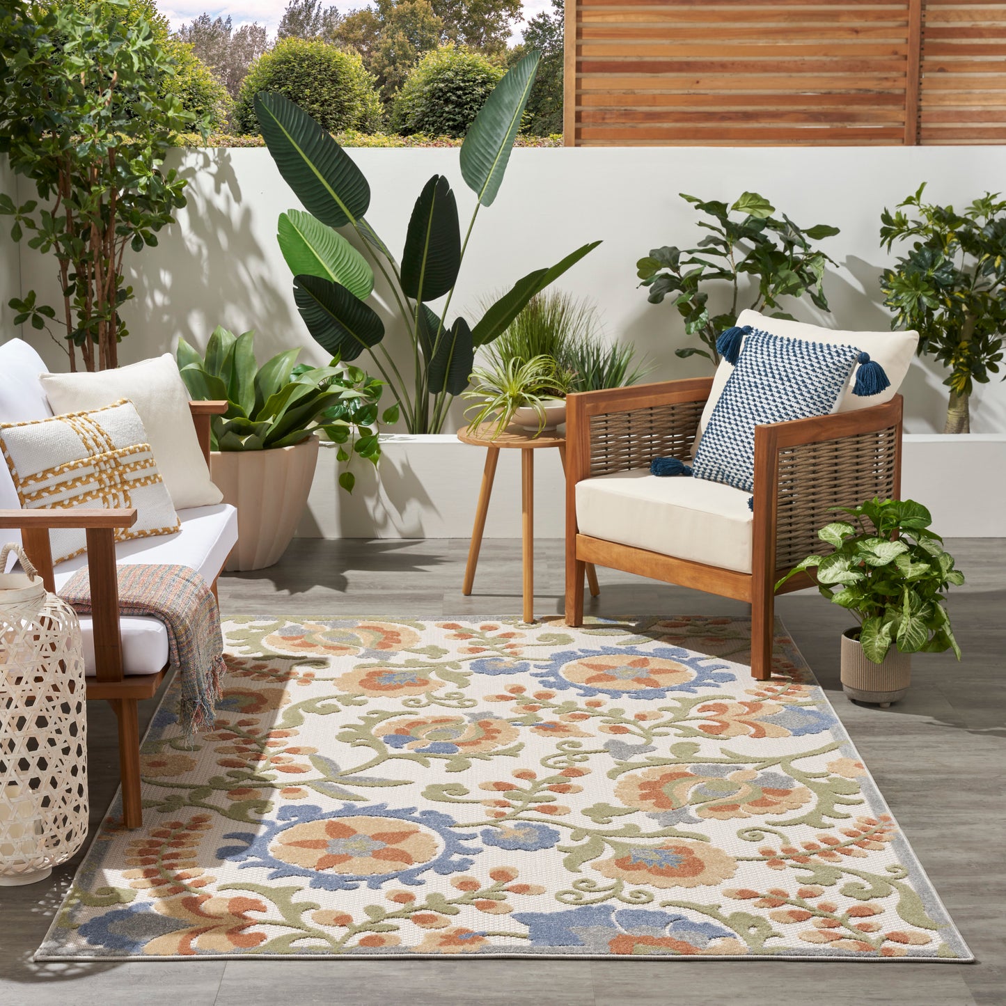Outdoor patio with wooden wicker chairs, cream cushions, colorful floral outdoor rug, potted tropical plants, sunlit garden.