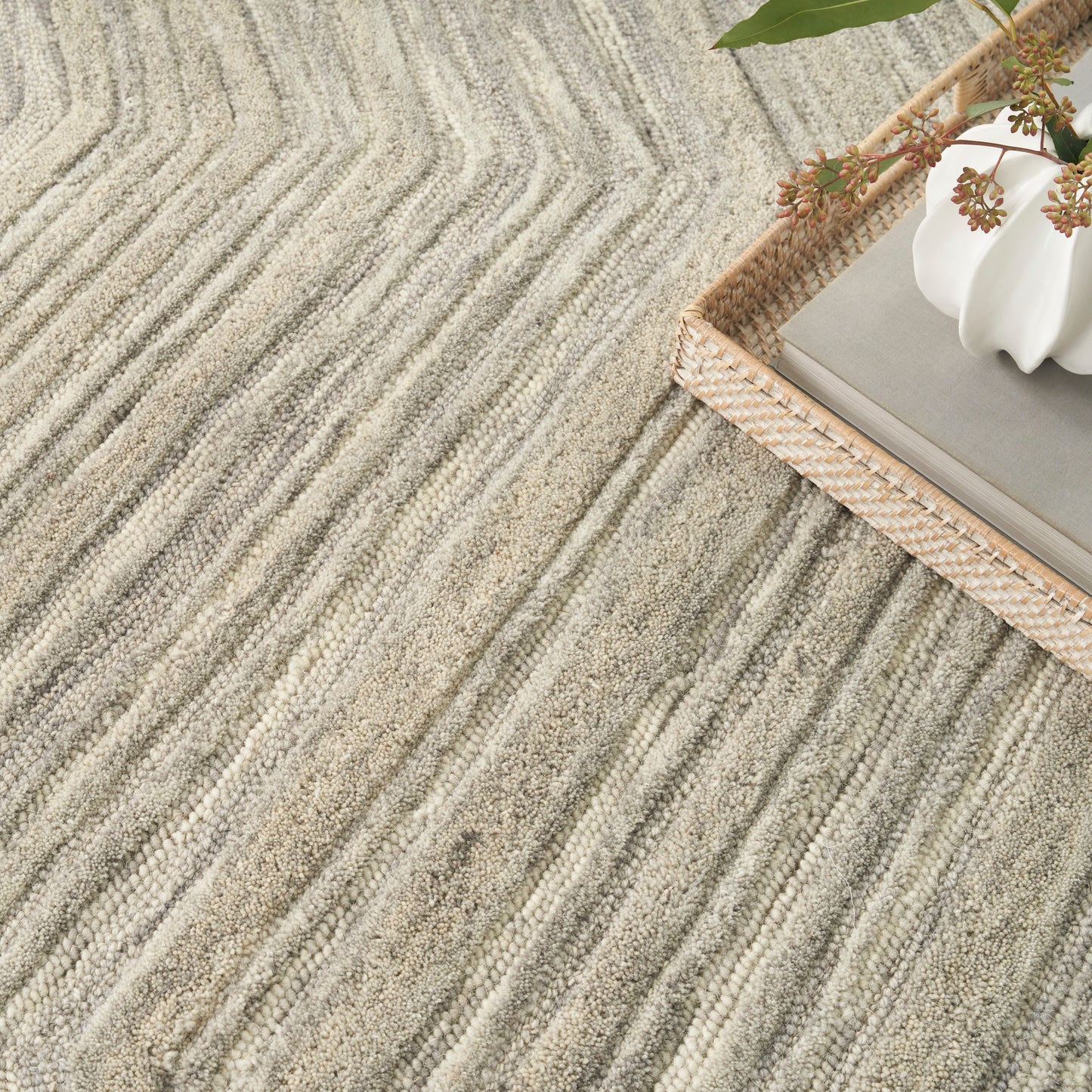 Beige striped textured rug with parallel grooves; a woven rattan basket holds a gray book & white vase with greenery.