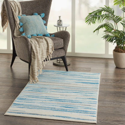 Gray rug with blue striped pattern, beige fringe throw, blue pom-pom pillow, and a potted plant by a sunny window.