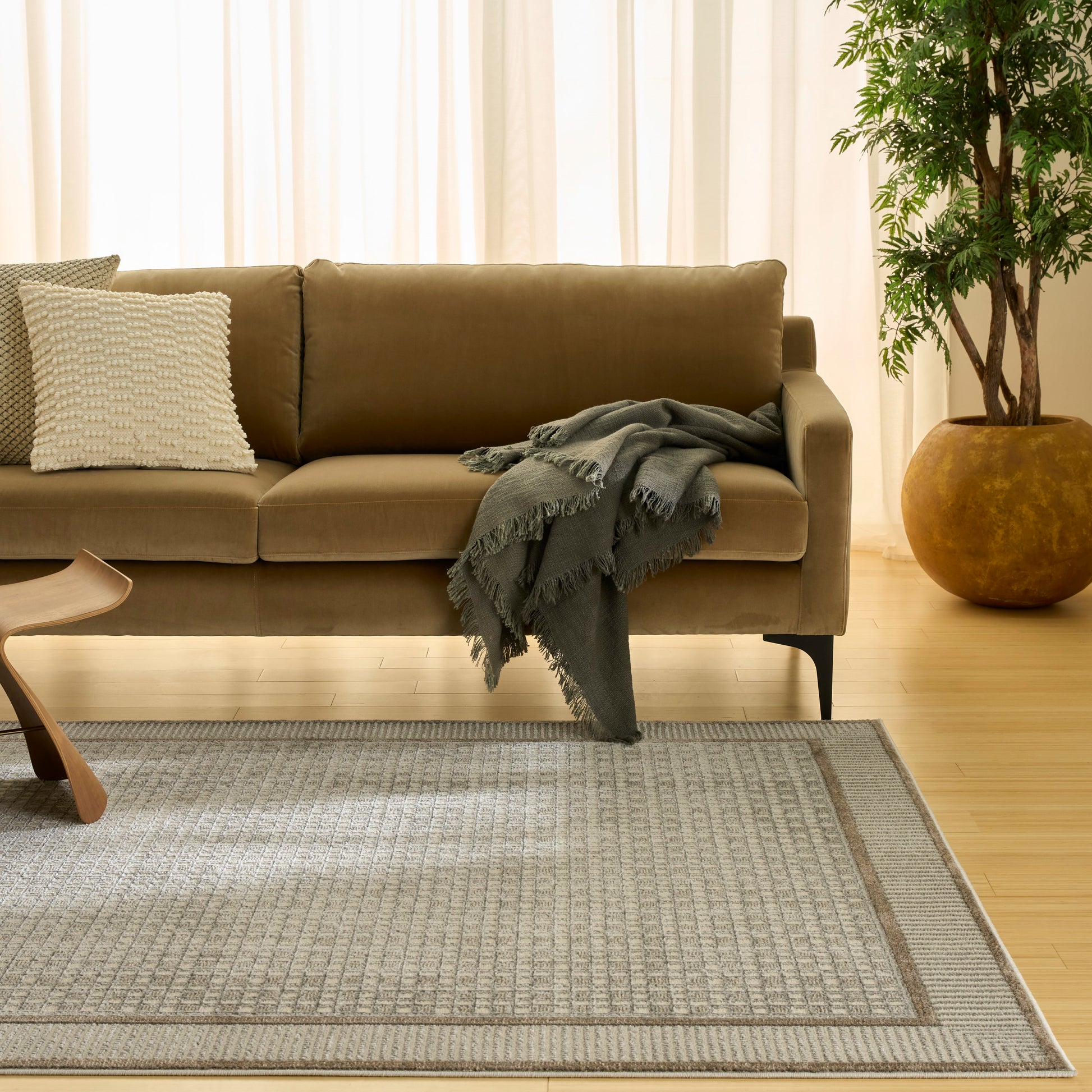 Camel sofa with white textured pillow and gray fringed throw on a woven rug; wooden chair, potted plant, beige curtains.