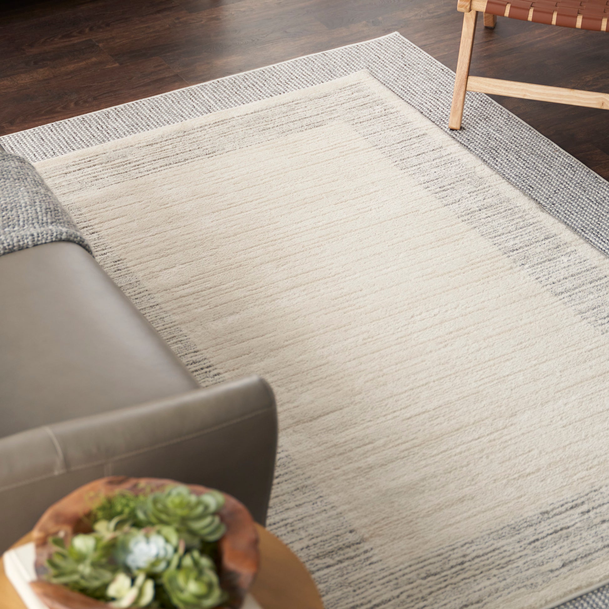 Rectangular rug with a light cream center and gray border, textured weave, in a modern living room beside a gray sofa.