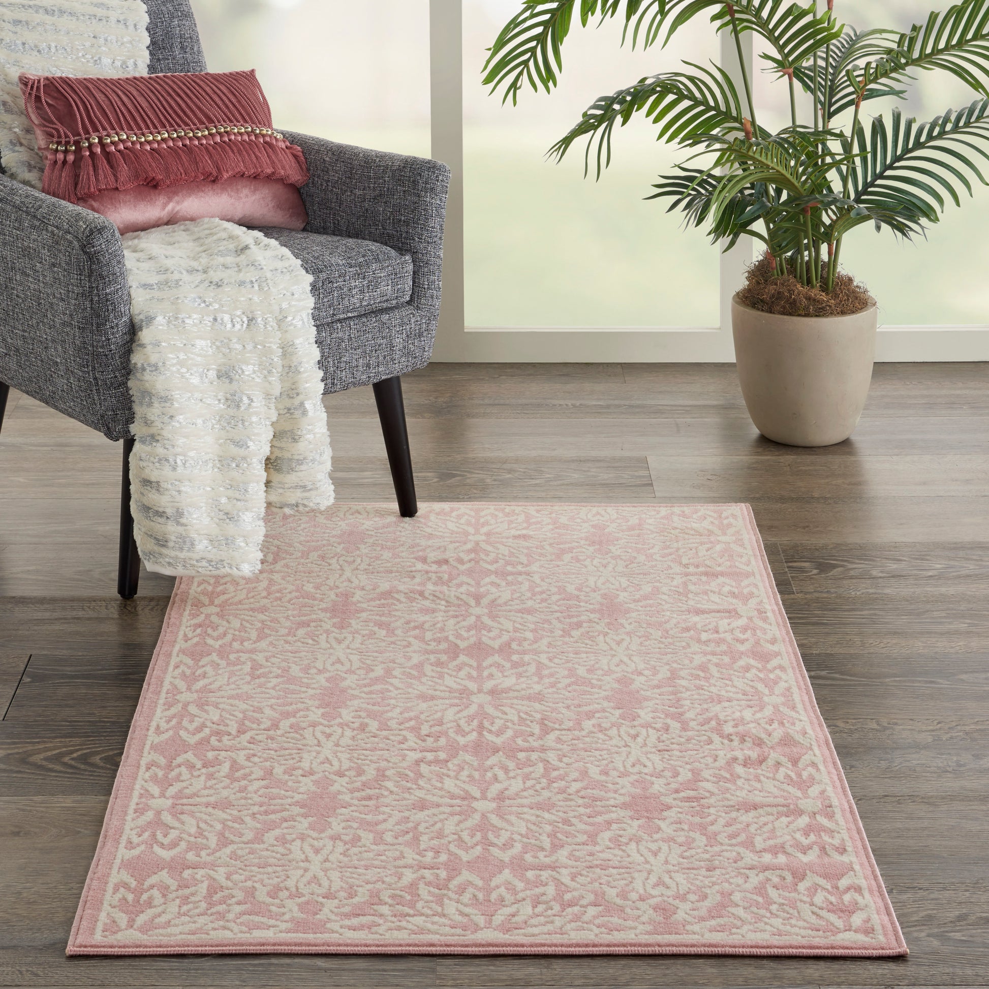 Pink patterned area rug on dark wood floor beside a grey armchair with a white throw and pink pillow, plant by the window.