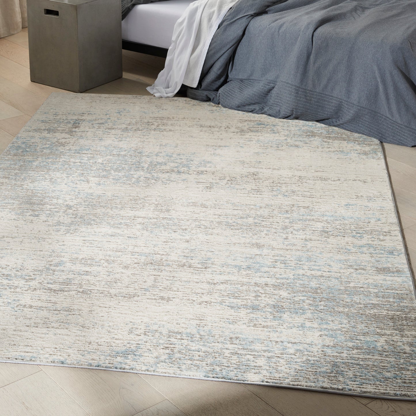 Rectangular abstract beige and light blue area rug in a modern bedroom beside a bed with gray-blue bedding.