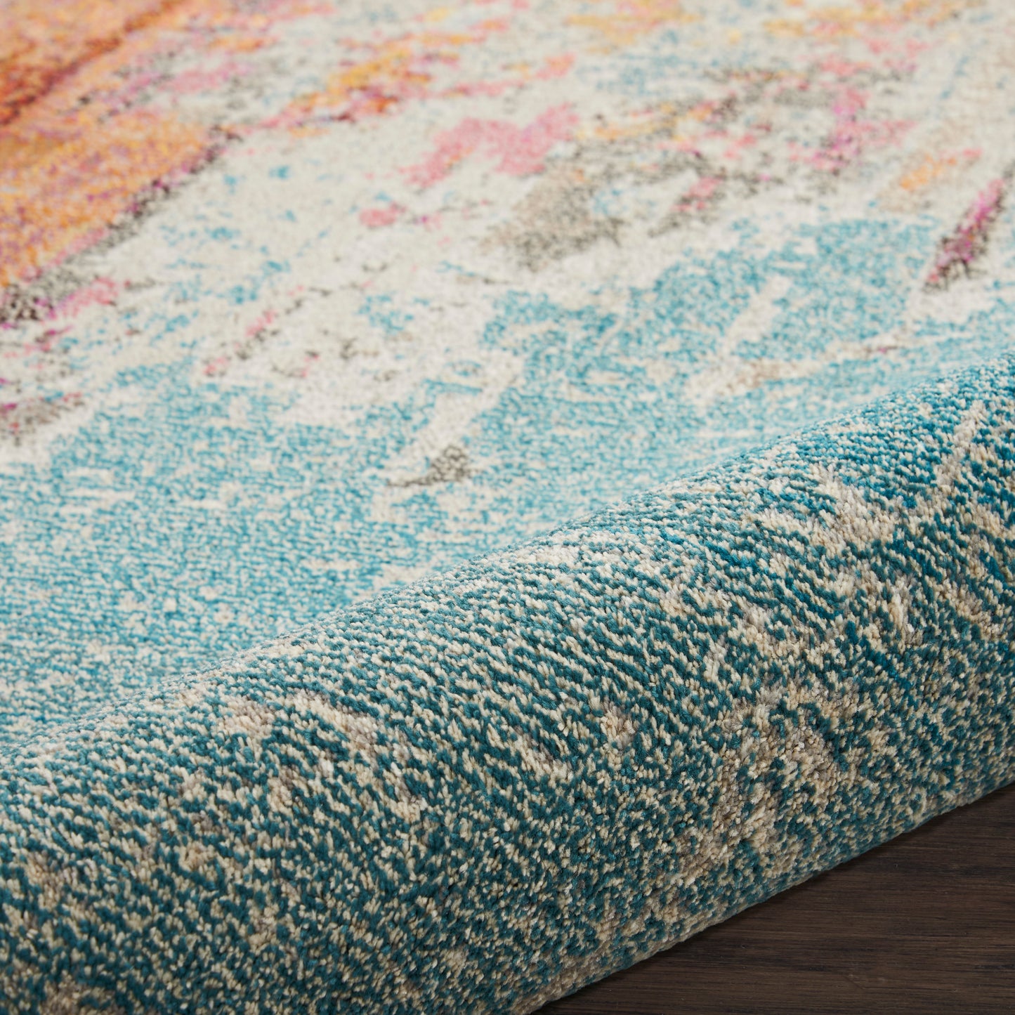 Close-up of a colorful abstract rug with teal edge and peach, pink, blue accents on dark wood floor.