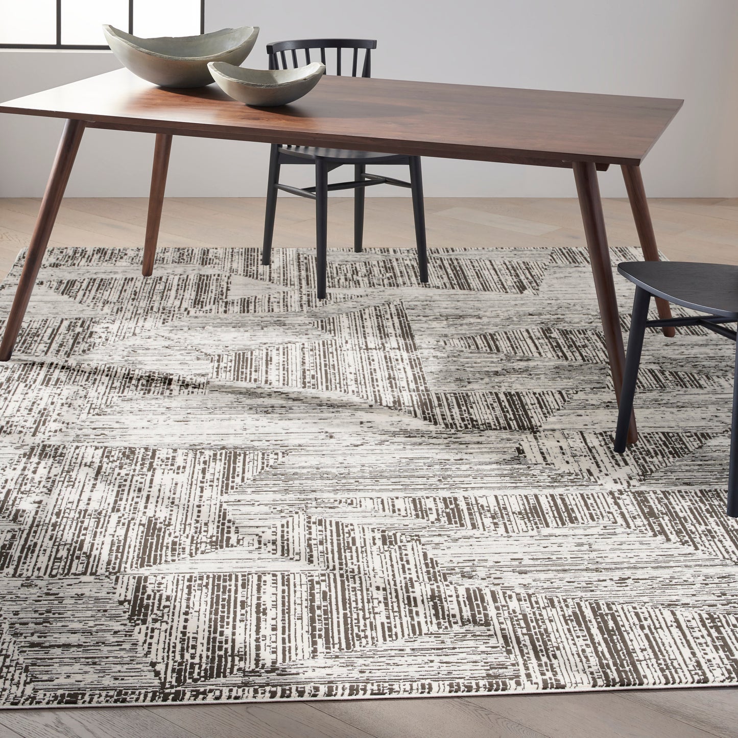 Mid-century walnut dining table with tapered legs, gray ceramic bowls & a black chair over a black-and-white rug.