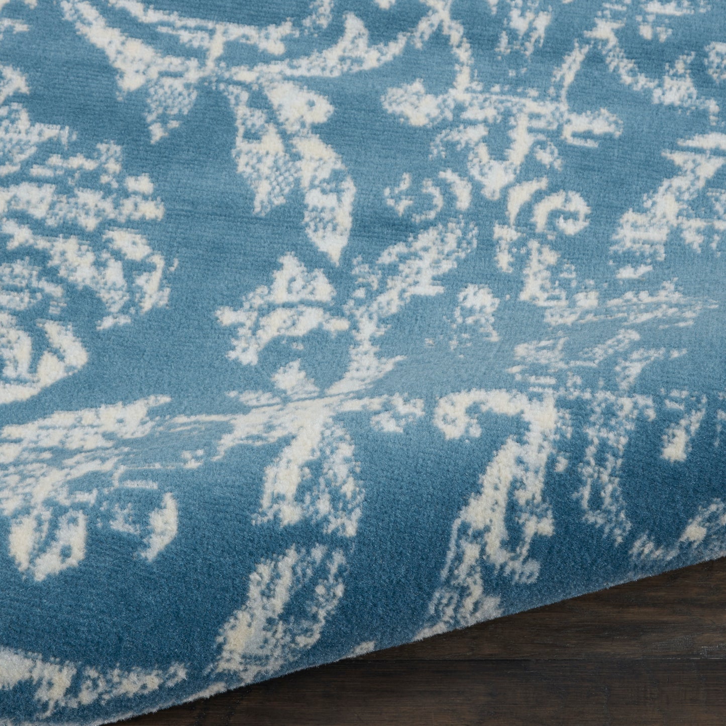 Close-up of a blue woven rug with a white distressed floral pattern on a dark wooden floor.