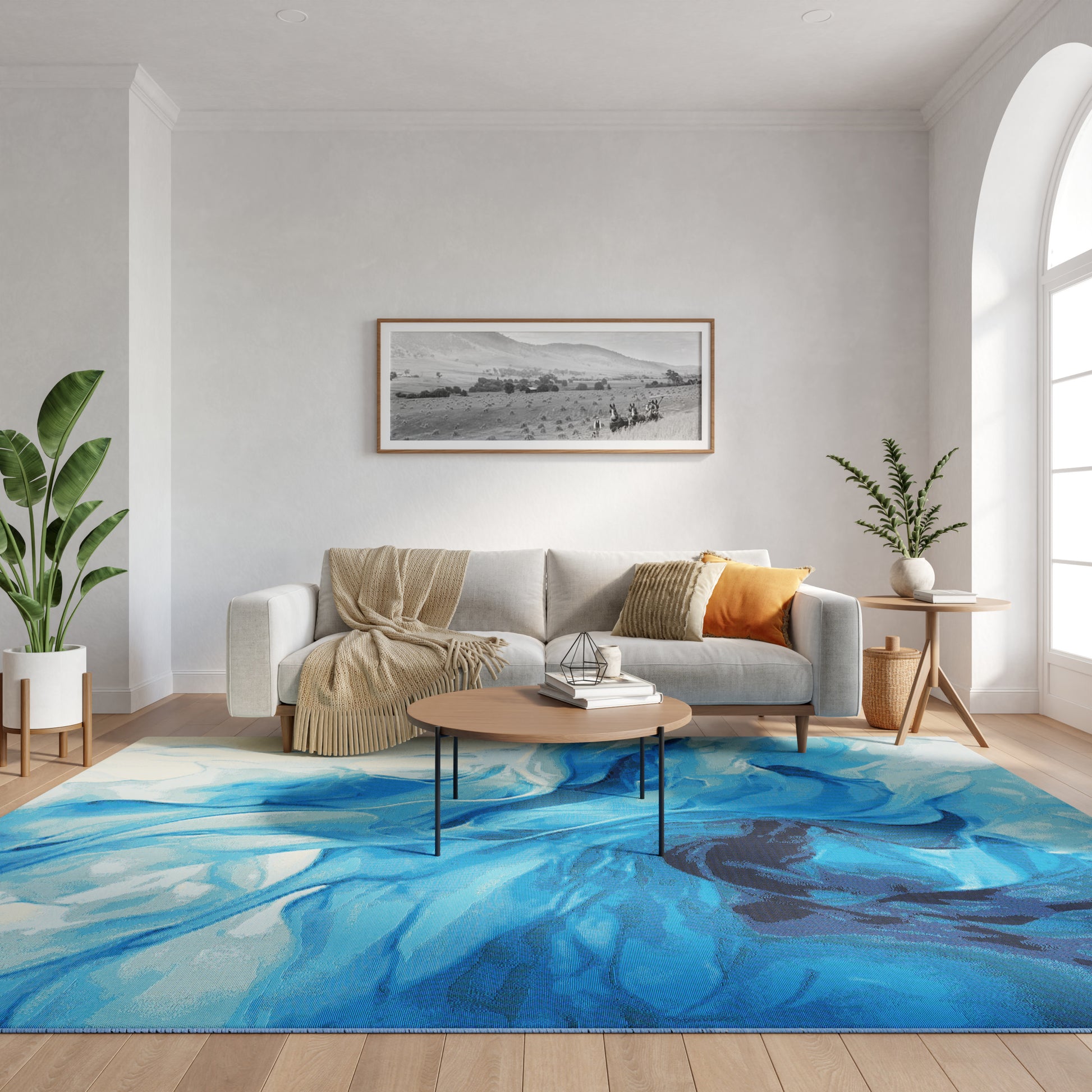 Light-filled living room with gray sofa, beige throw, orange pillow, blue abstract rug, round wood coffee table, and greenery