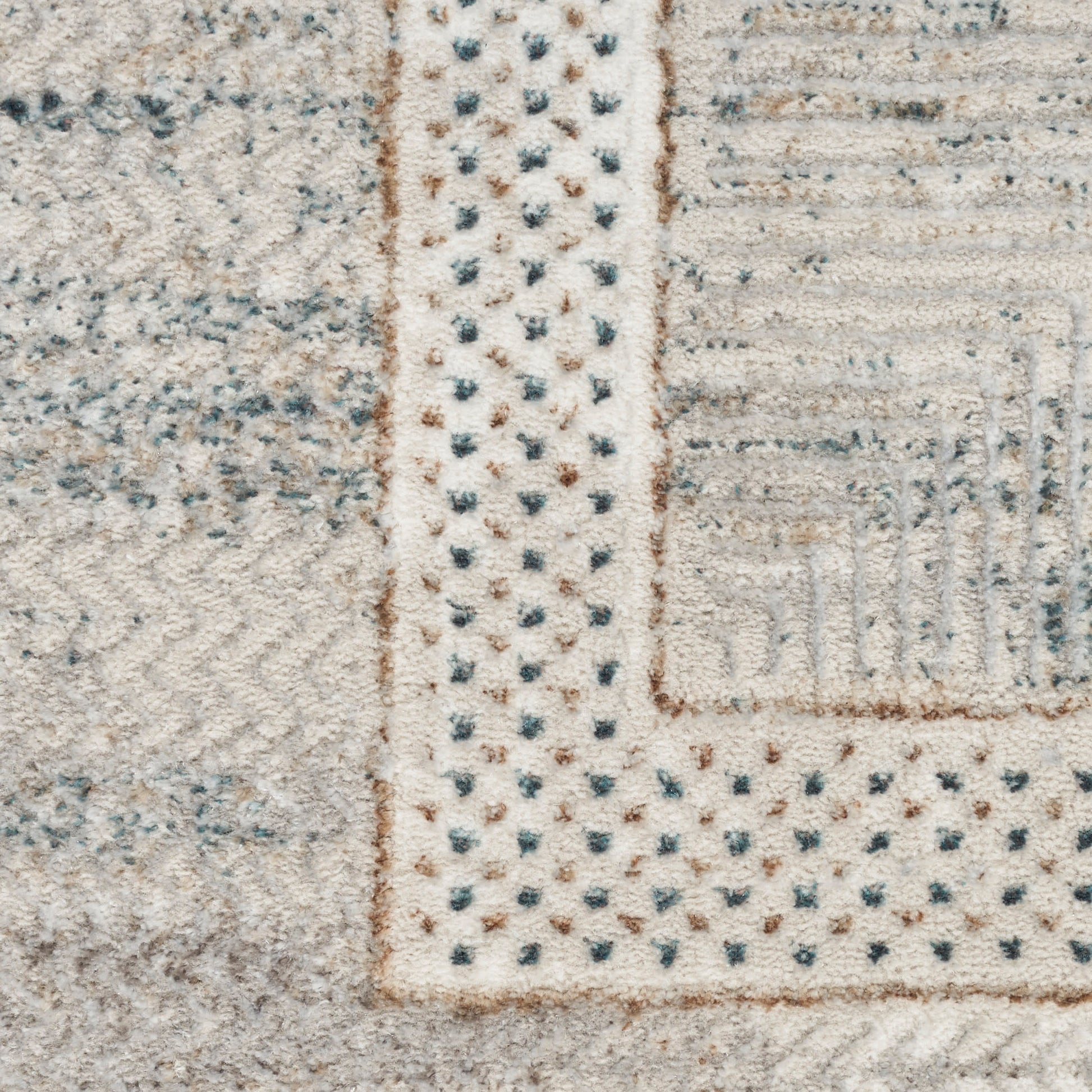 Close-up of a woven pile rug in light beige with a dotted border and patchwork geometric blocks in cream, grey, and blue.