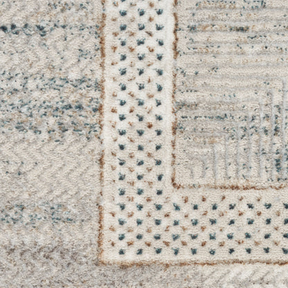 Close-up of a woven pile rug in light beige with a dotted border and patchwork geometric blocks in cream, grey, and blue.