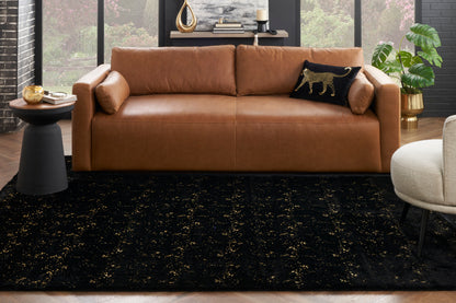 Caramel leather 3-seat sofa with squared arms on a black & gold-speckled rug; modern living room with plant & round side table.